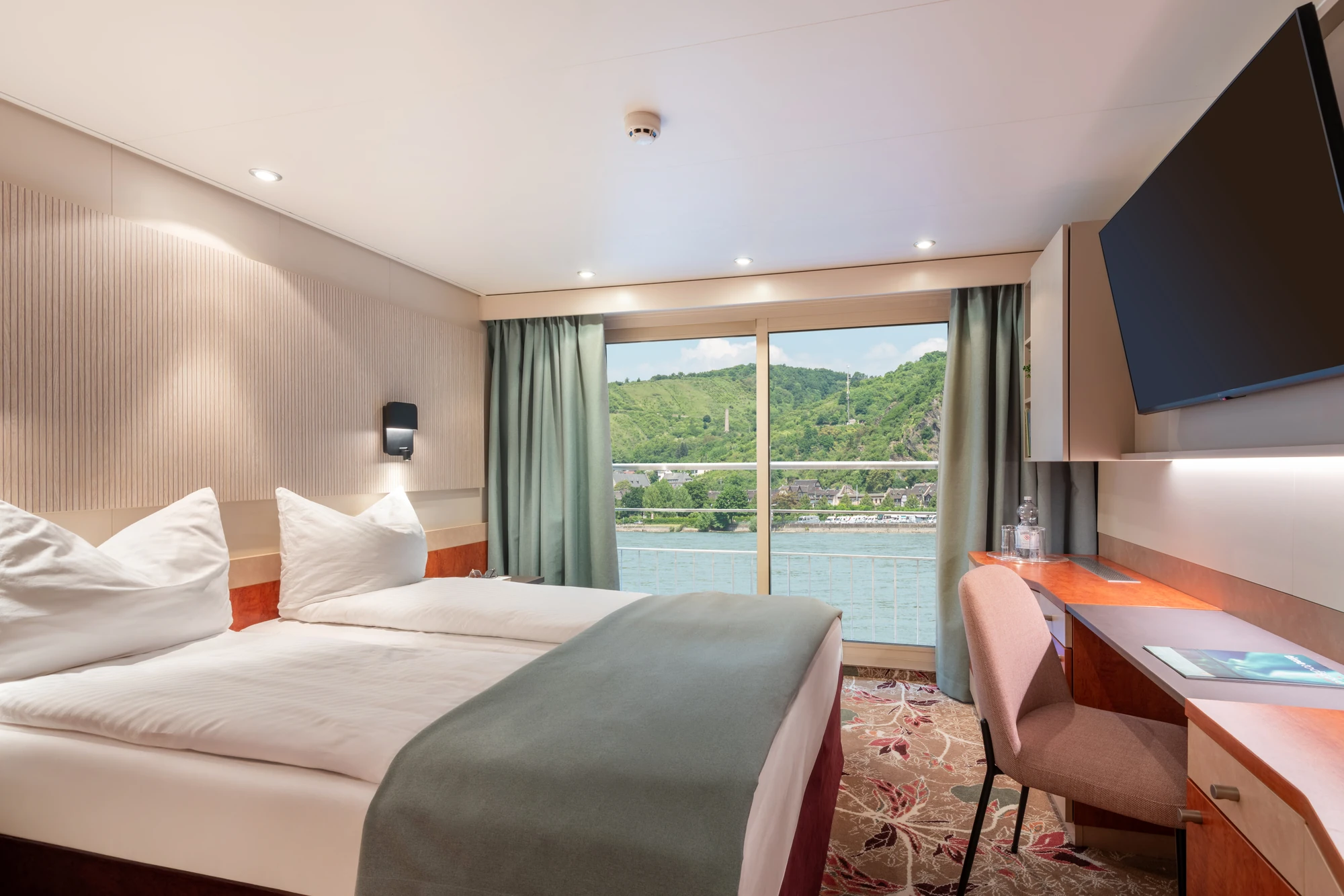 Zweibettzimmer mit zwei Einzelbetten, grünem Bettläufer, Fenster mit Blick auf Fluss und bewaldete Hügel, Schreibtisch mit Stuhl und Wandfernseher.