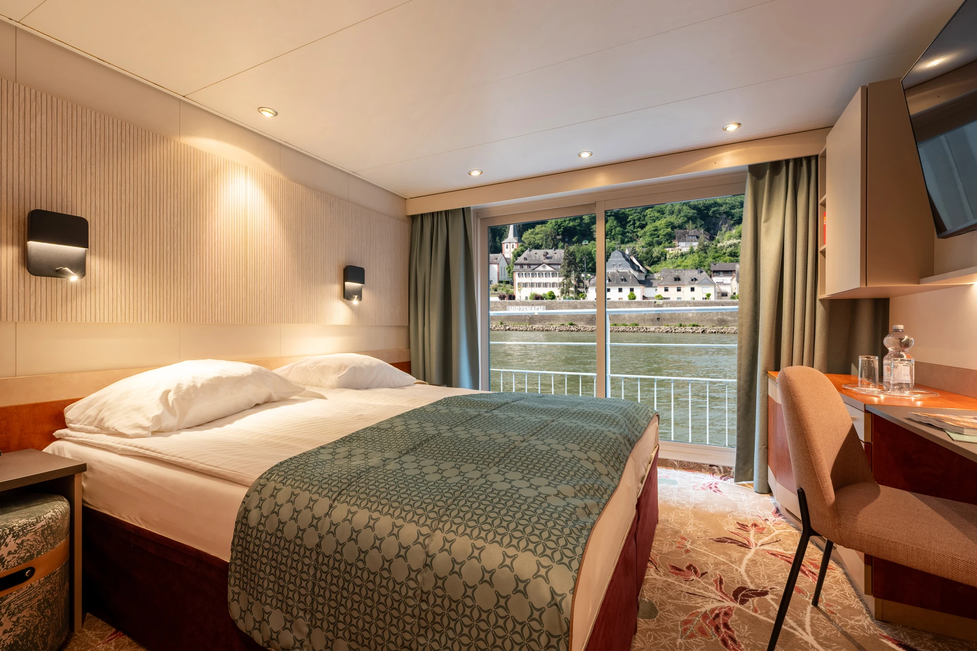 Doppelbett mit gemustertem Überwurf in Kabine mit Fenster und Blick auf Fluss und Häuser, Schreibtisch mit Stuhl und Fernseher an der Wand.