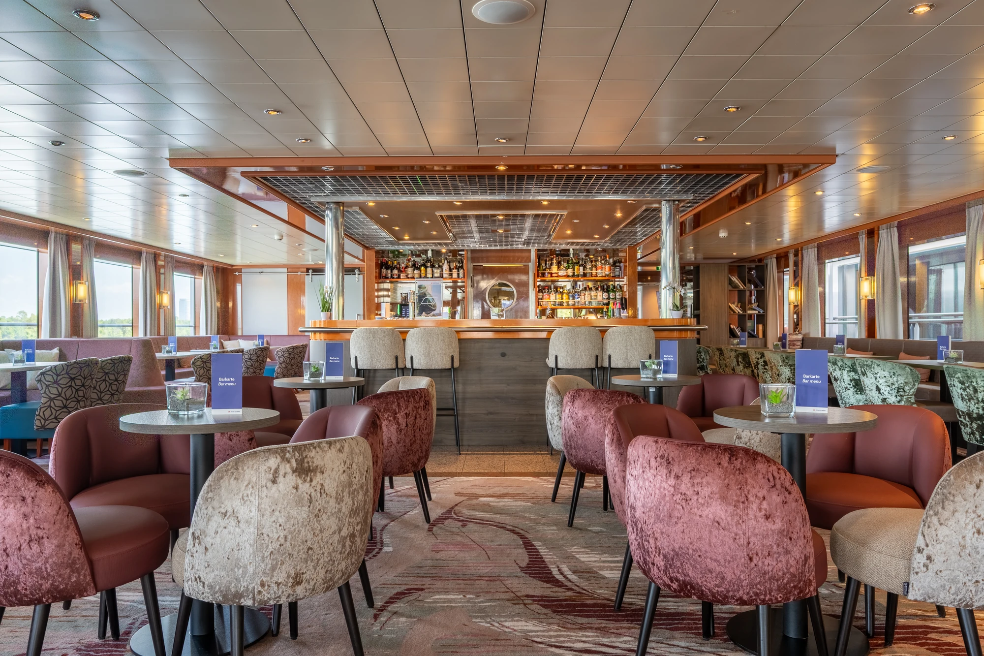 Moderne Bar mit gepolsterten Sesseln in verschiedenen Farben, Tischen mit Menükarten und einer beleuchteten Bar im Hintergrund.