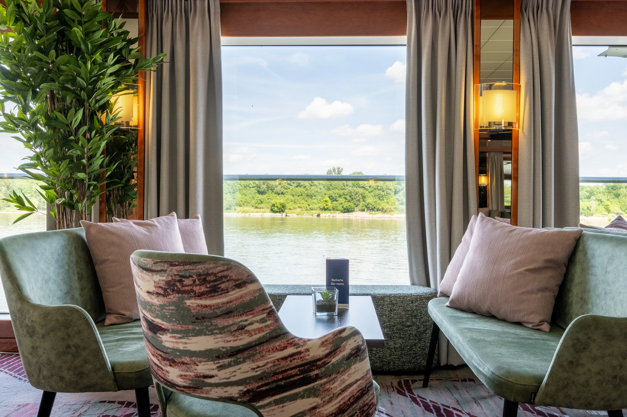 Panoramafenster mit Blick auf Fluss, davor zwei grüne Sofas mit Kissen, ein gemusterter Sessel und ein kleiner Tisch mit Karte.
