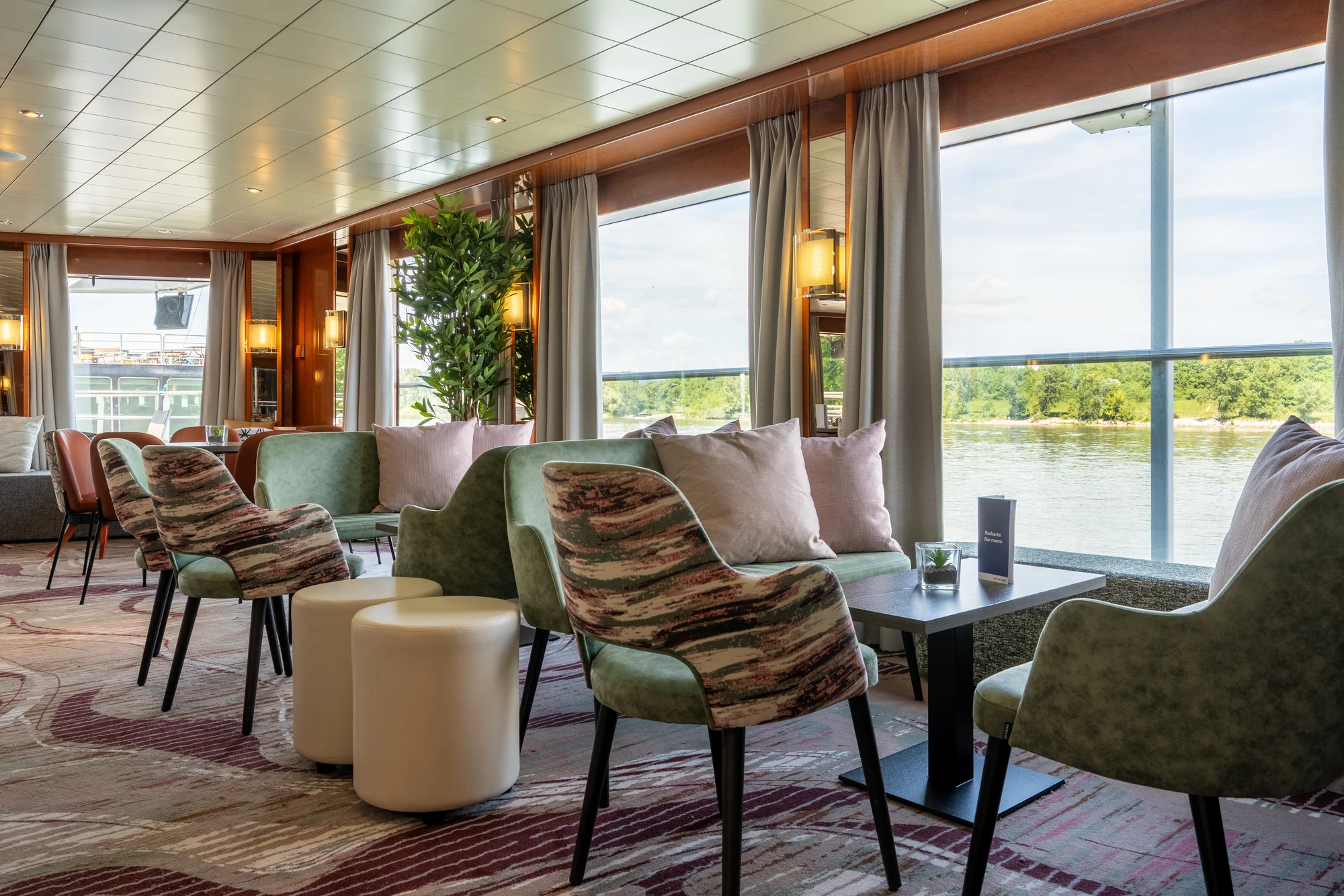 Panoramafenster mit Blick auf Fluss, davor Sitzgruppe mit grünen Sesseln, gemusterten Stühlen, Kissen und kleinen Tischen.