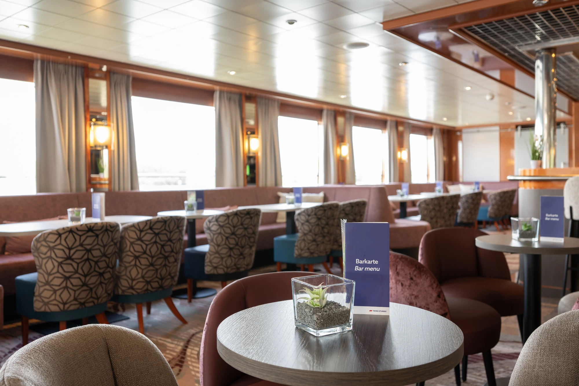 Panoramablick in einen Salon mit mehreren Tischen, gepolsterten Stühlen und großen Fenstern mit Vorhängen.