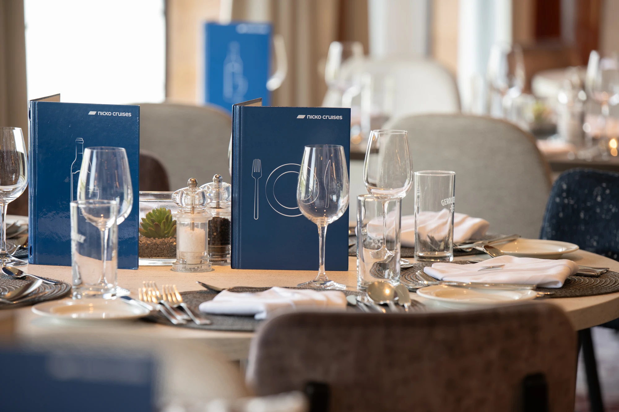 Runder Tisch im Restaurant mit blauem Menü von Fred. Olsen Cruise Lines, Weingläsern, Wassergläsern, Besteck und weißen Servietten.