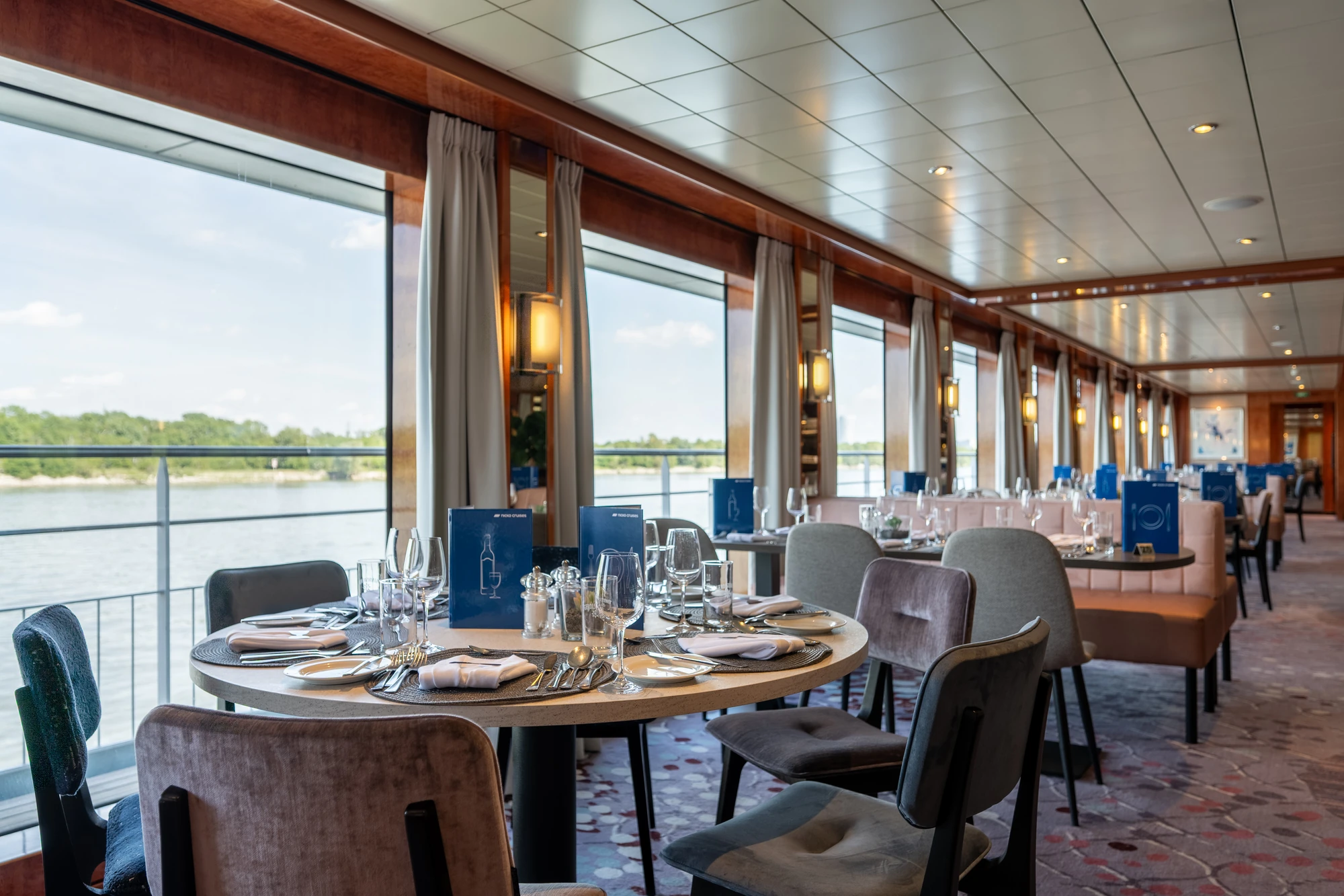 Runder Tisch im Restaurant mit blauem Menü von Fred. Olsen Cruise Lines, Weingläsern, Wassergläsern, Besteck und weißen Servietten.