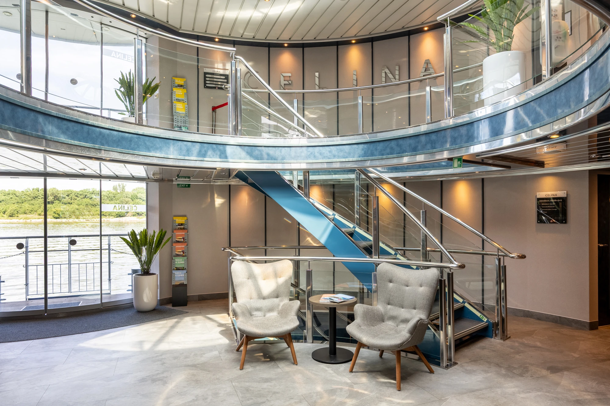 Helle Empfangshalle mit zwei grauen Sesseln, kleinem runden Tisch, blauen Treppenstufen und großen Fenstern mit Blick auf Fluss und Bäume.