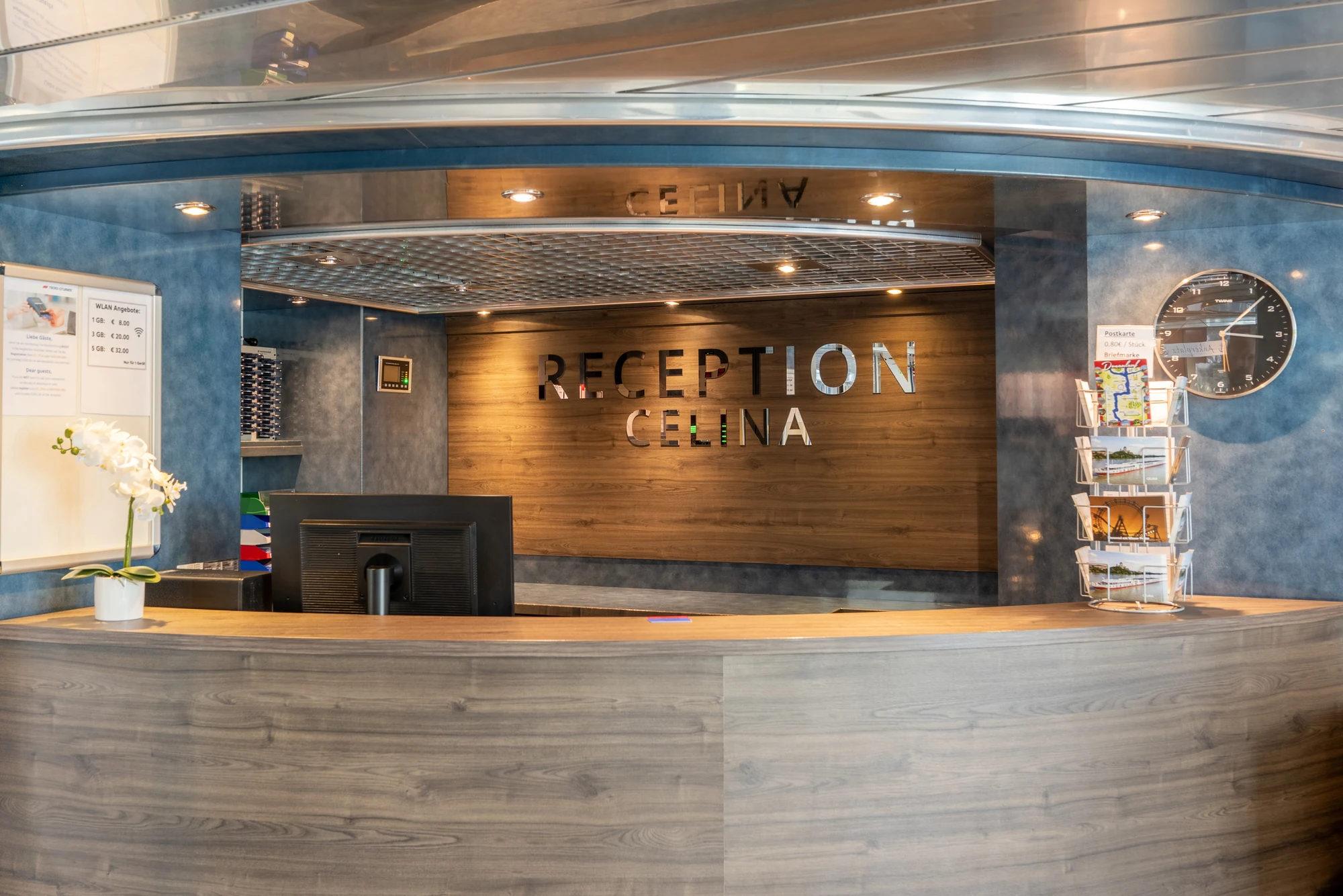 Rezeption mit Holzverkleidung, Schriftzug 'RECEPTION CELINA' an der Wand, Computerbildschirm, weiße Orchidee auf dem Tresen, Wanduhr und Prospektständer.