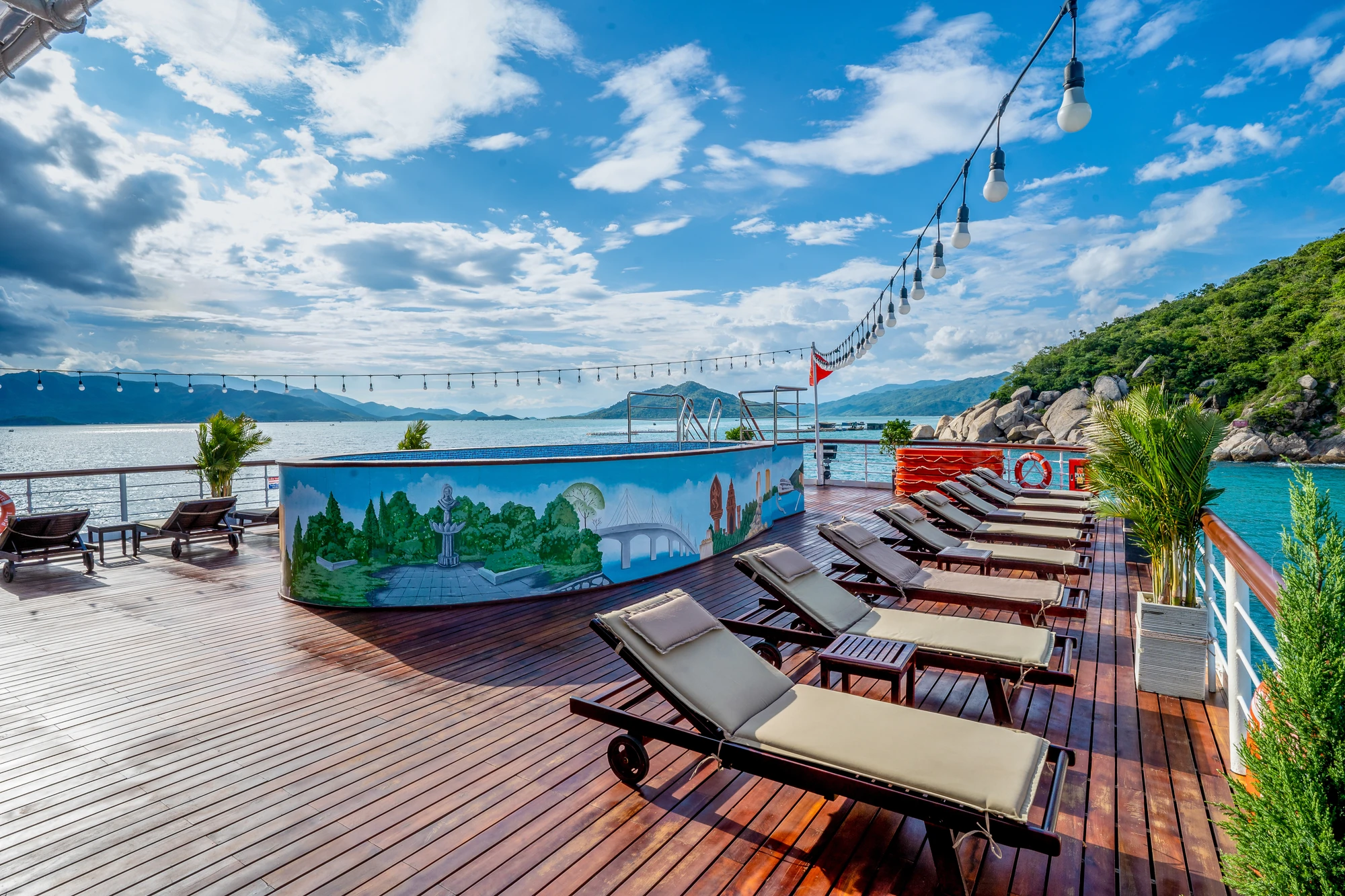 Holzdeck mit Liegen, einer gemalten Wand mit Landschaftsmotiv und einer Aussicht auf das Wasser. Lichterketten hängen über dem Deck.