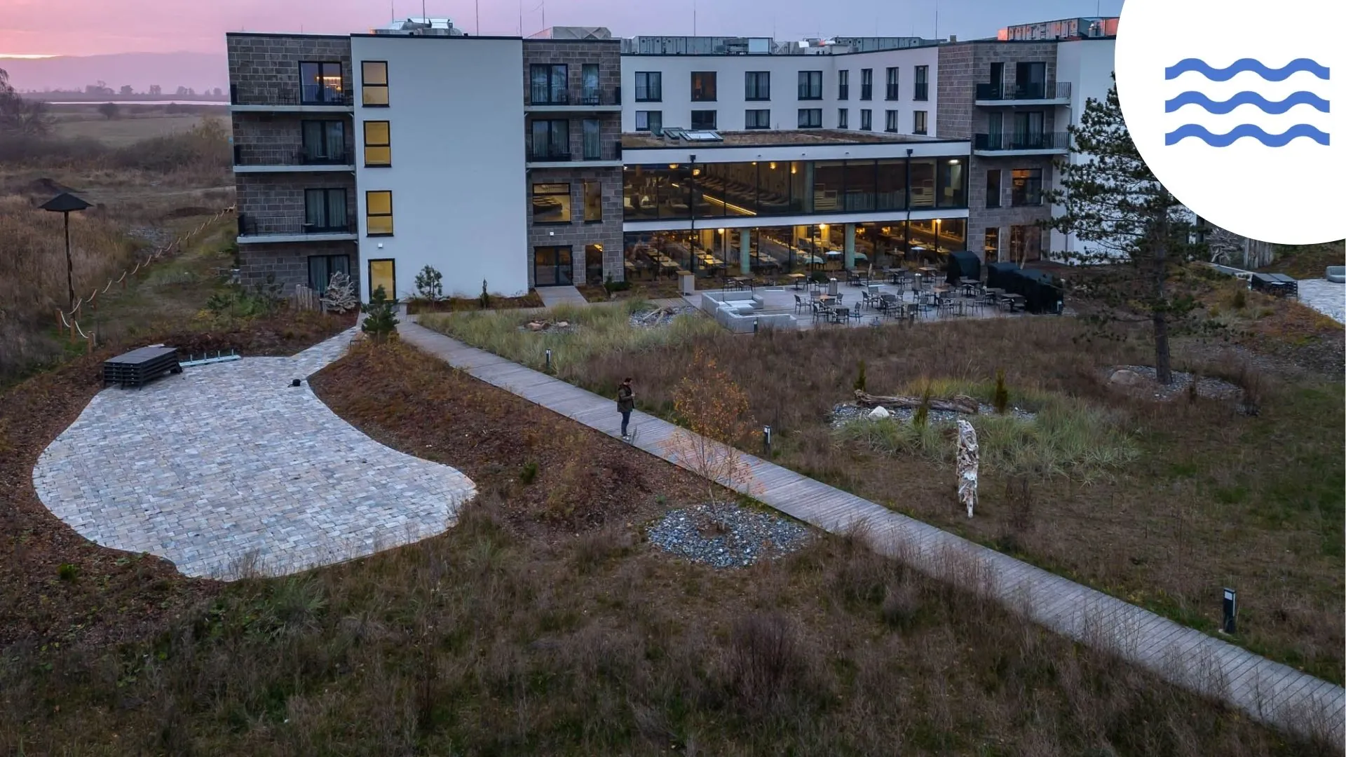 Santé Royale Rügen Außenansicht bei Sonnenuntergang: Das Gebäude mit weißen Wänden und Naturstein steht inmitten der Natur. Ein Holzweg führt zum Hotel. Im Hintergrund kann man einen kleinen Teil der Ostsee erkennen.