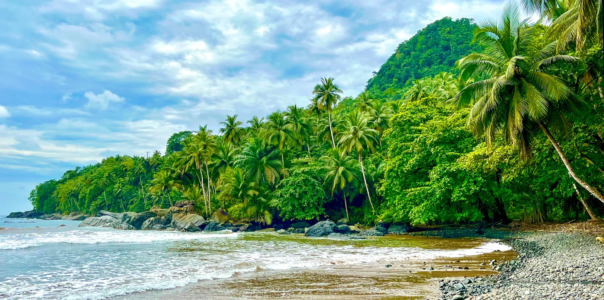 Strand mit Sand, Felsen und Palmen vor bewaldeten Hügeln unter bewölktem Himmel.