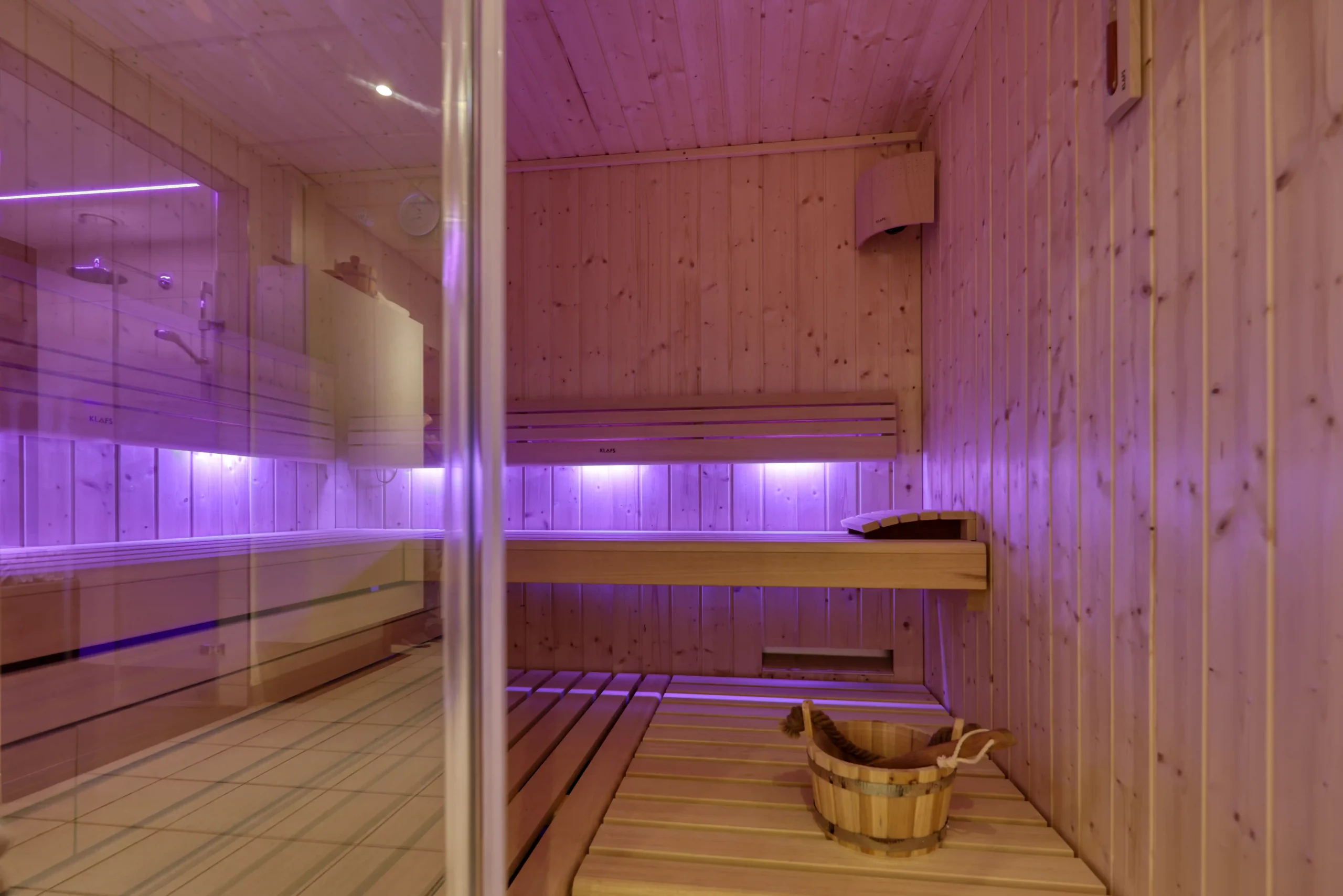 Saunabereich mit Holzbänken, Holzwänden, einem Holzeimer mit Schöpfkelle und violettem Licht hinter einer Glastür
