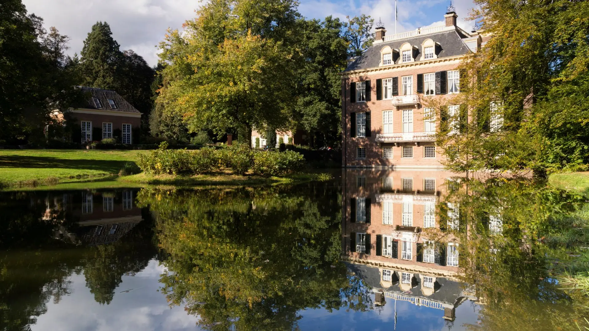 Schloss Rosendael in Arnhem mit mehrstöckigem Backsteingebäude, umgeben von Bäumen und spiegelndem Teich im Vordergrund.