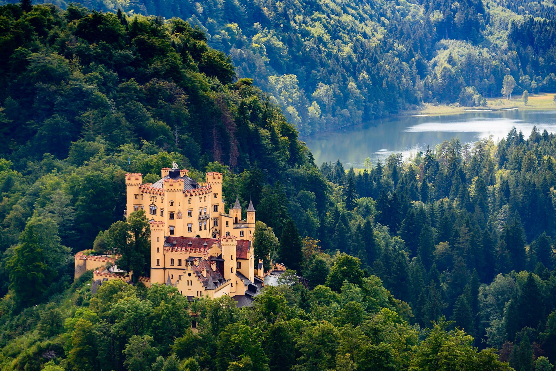 Das Bild zeigt Schloss Hohenschwangau, malerisch gelegen inmitten dichter Wälder, umgeben von der beeindruckenden Naturkulisse der bayerischen Alpen. Das gelb getünchte Schloss erhebt sich majestätisch über den grünen Baumwipfeln, während im Hintergrund ein ruhiger See glitzert. Diese idyllische und historische Szenerie unterstreicht die romantische Atmosphäre, die das Schloss ausstrahlt, und macht es zu einem beliebten Reiseziel.