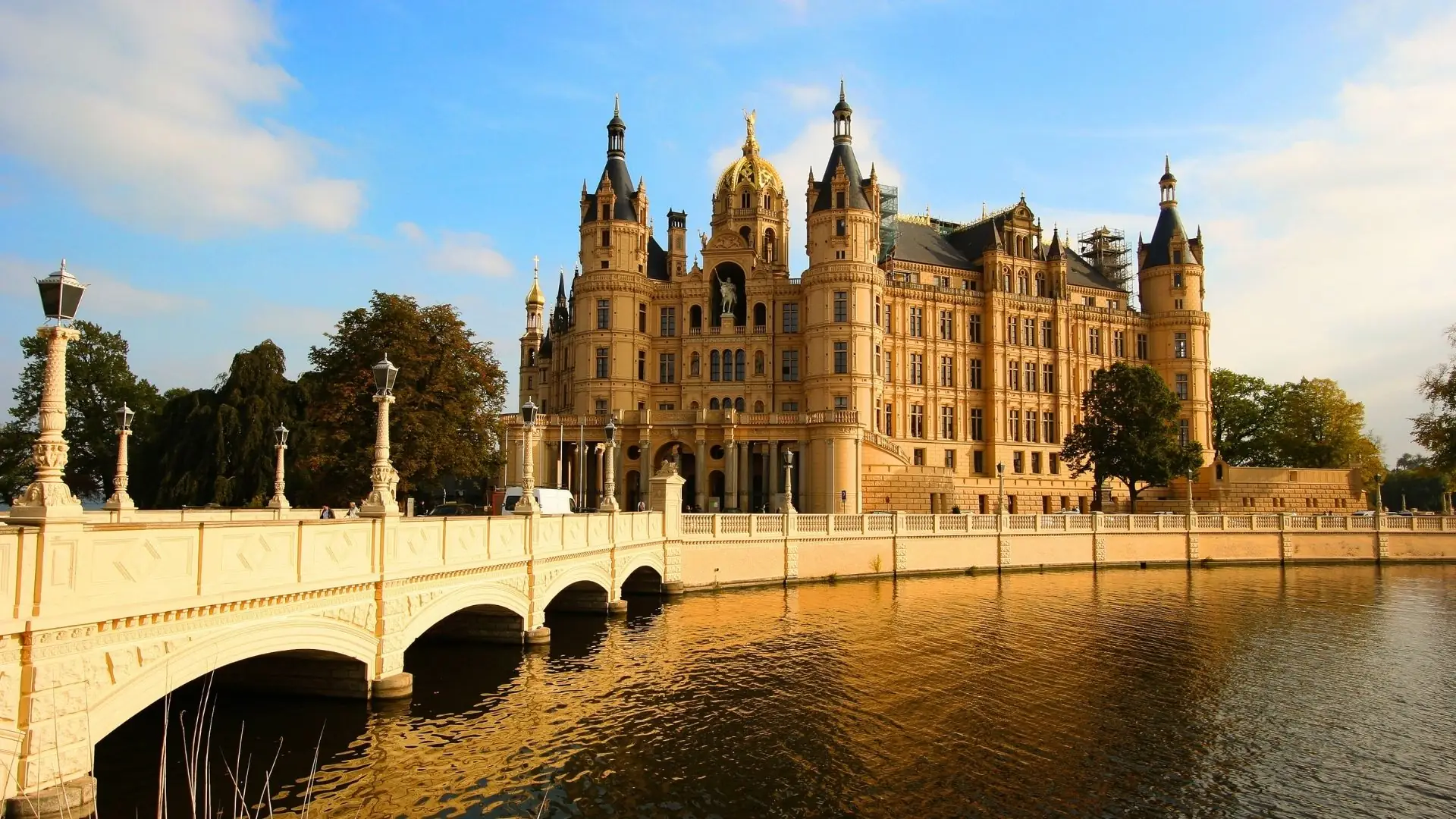 Schloss Schwerin mit goldenen Türmen und Brücke, umgeben von einem malerischen Gewässer bei Sonnenlicht.