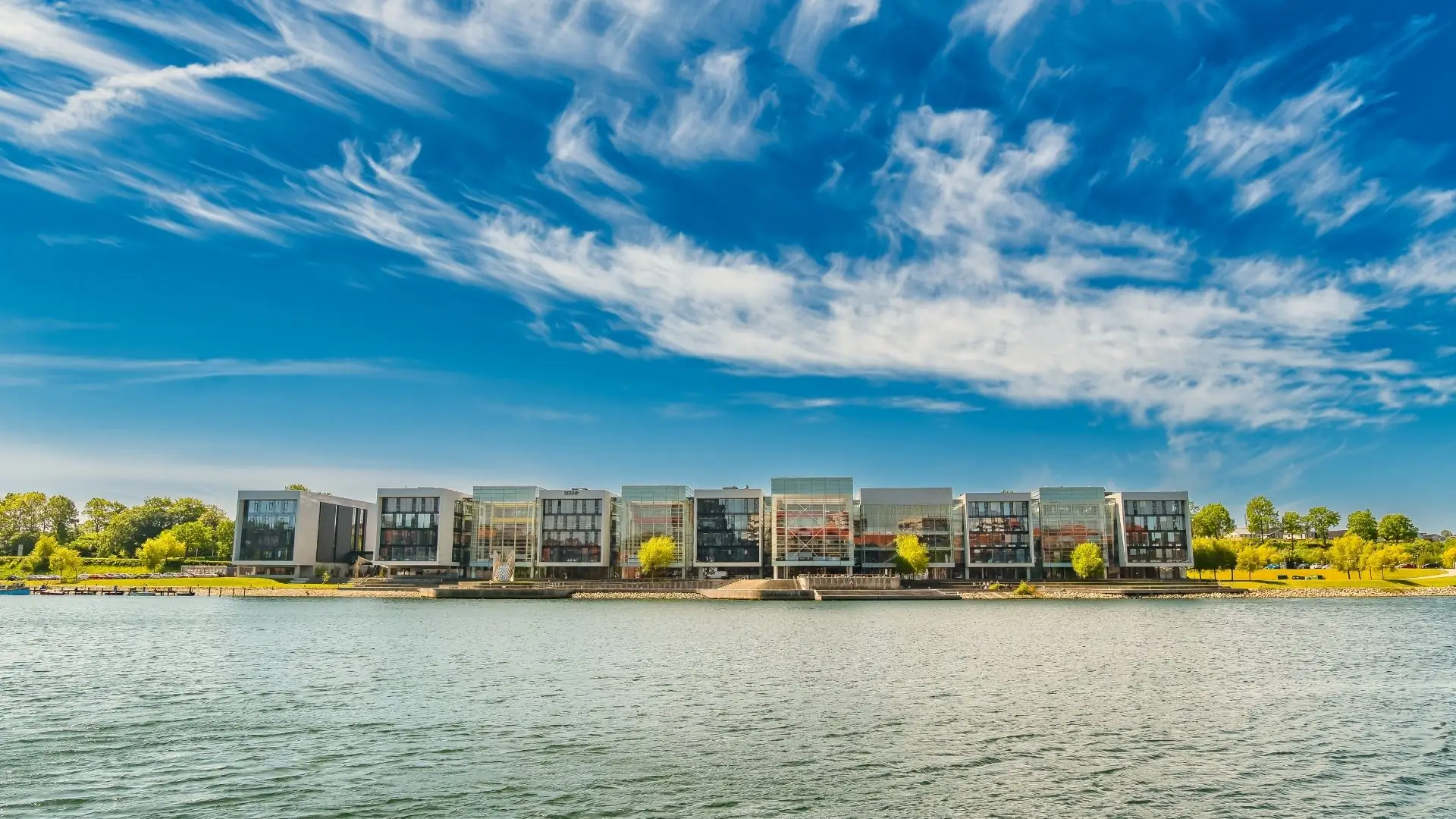 Reihe moderner Wohngebäude mit großen Fenstern am Ufer, davor Wasser, im Hintergrund Bäume und bewölkter Himmel in Sonderburg, Dänemark.