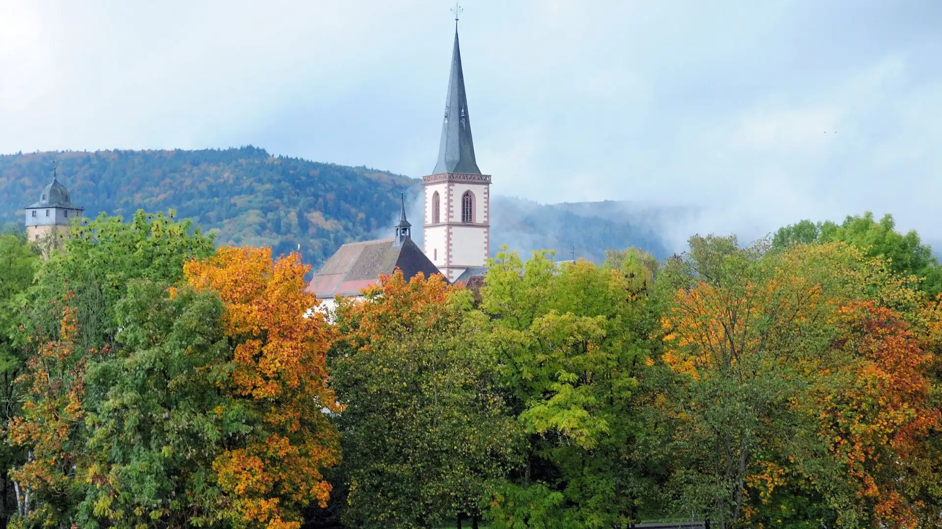 Kirchturm der Stadtkirche Lohr am Main ragt über herbstlich gefärbte Bäume vor bewaldeten Hügeln mit leichtem Nebel