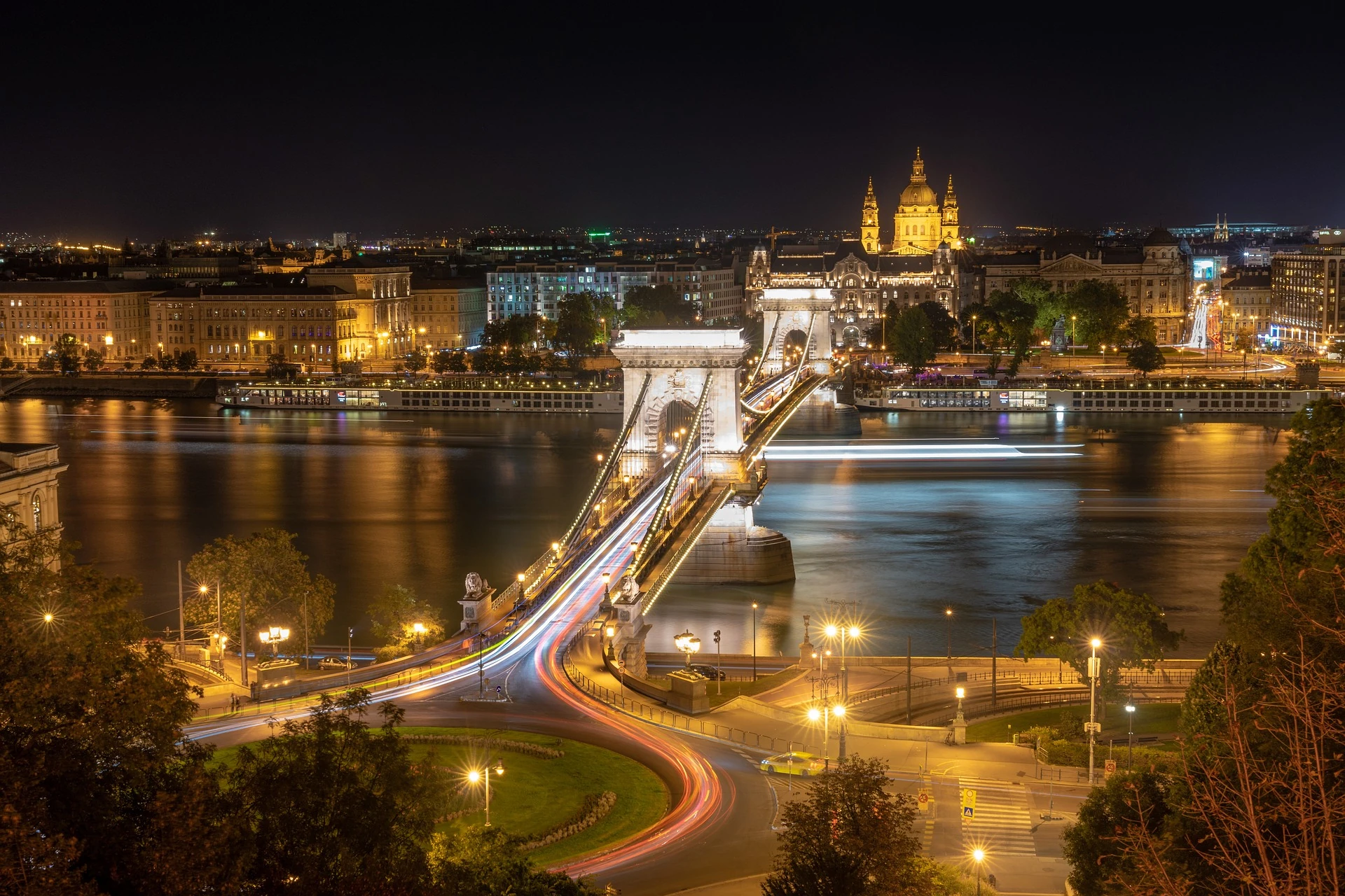 Städtereise Budapest zeigt die beleuchtete Kettenbrücke bei Nacht, die über die Donau führt, mit der eindrucksvoll erleuchteten St.-Stephans-Basilika und den historischen Gebäuden im Hintergrund.