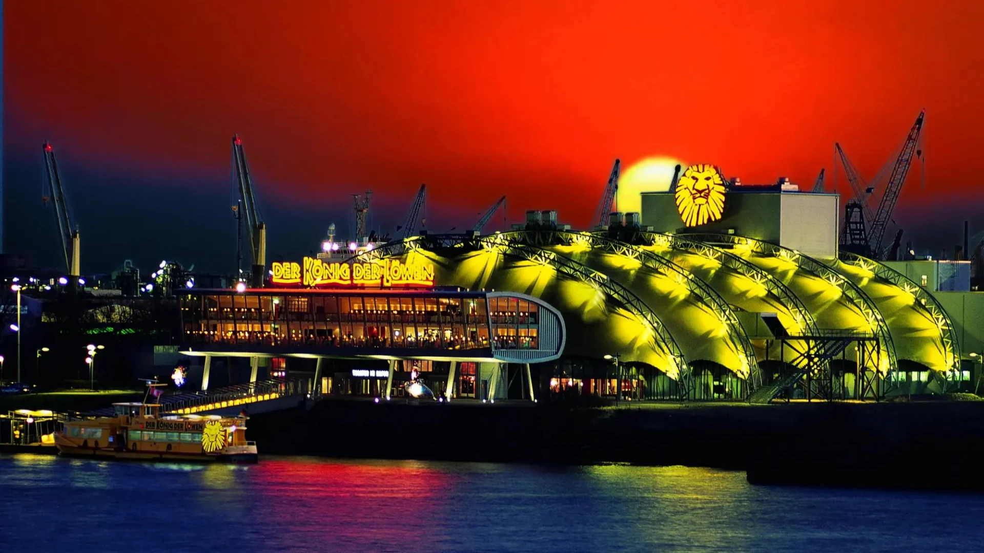 Die lebhafte Szene zeigt das Stage Theater am Hafen „Der König der Löwen“, das vor einem dramatischen roten und blauen Sonnenuntergang beleuchtet wird und sich im Wasser spiegelt.