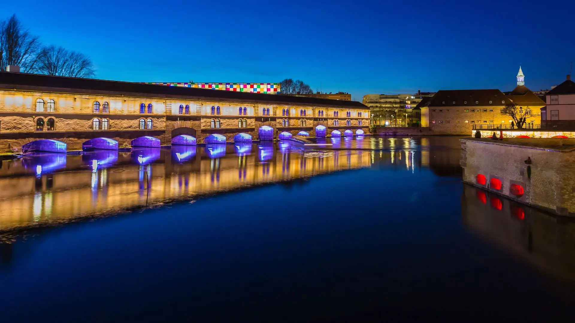 Barrage Vauban in Straßburg bei Nacht, beleuchtete Steinbrücke mit blauem Licht unter den Bögen und roten Lichtern am Ufer, Spiegelung im Wasser.