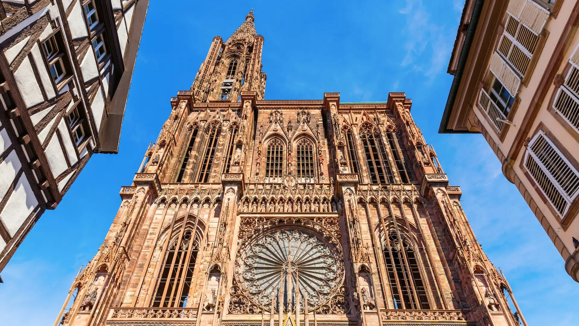 Fassade des Straßburger Liebfrauenmünsters mit gotischem Turm, Rosettenfenster und Fachwerkhäusern links und rechts, blauer Himmel im Hintergrund.