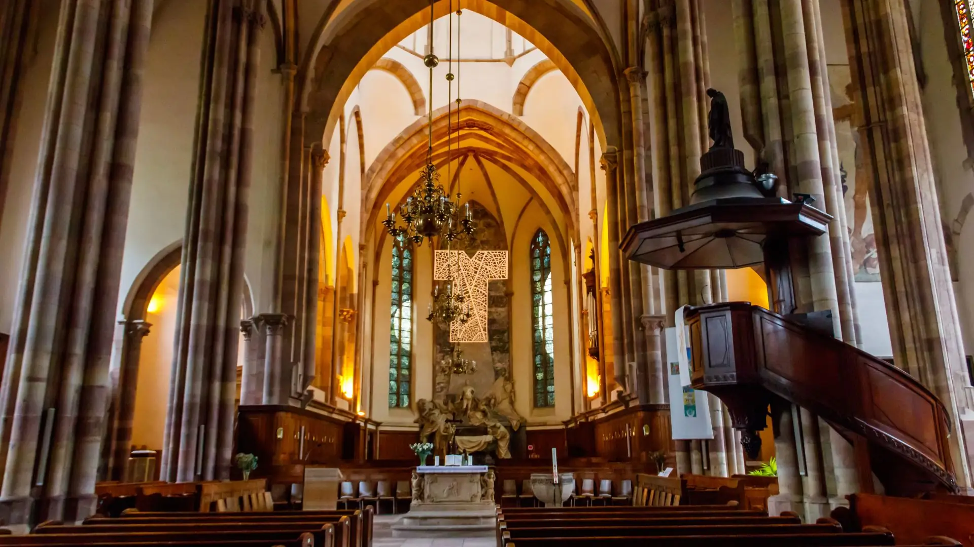 Innenraum der Thomaskirche Straßburg mit gotischen Spitzbögen, hölzernen Kirchenbänken, Altar und hängendem Kronleuchter.