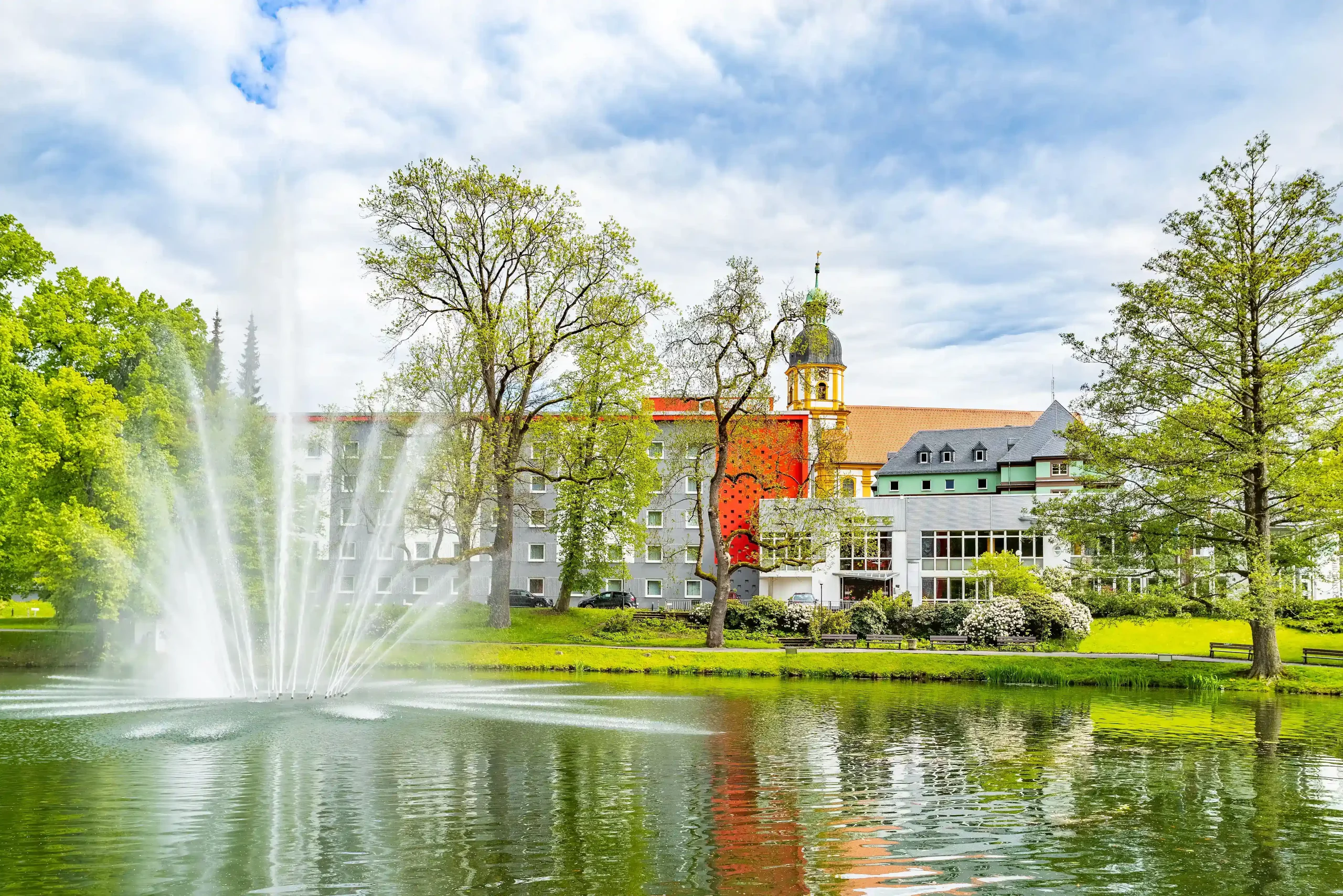 Hotelgebäude des ACHAT Hotel Suhl mit angrenzendem Teich und Springbrunnen, umgeben von Bäumen und Grünflächen