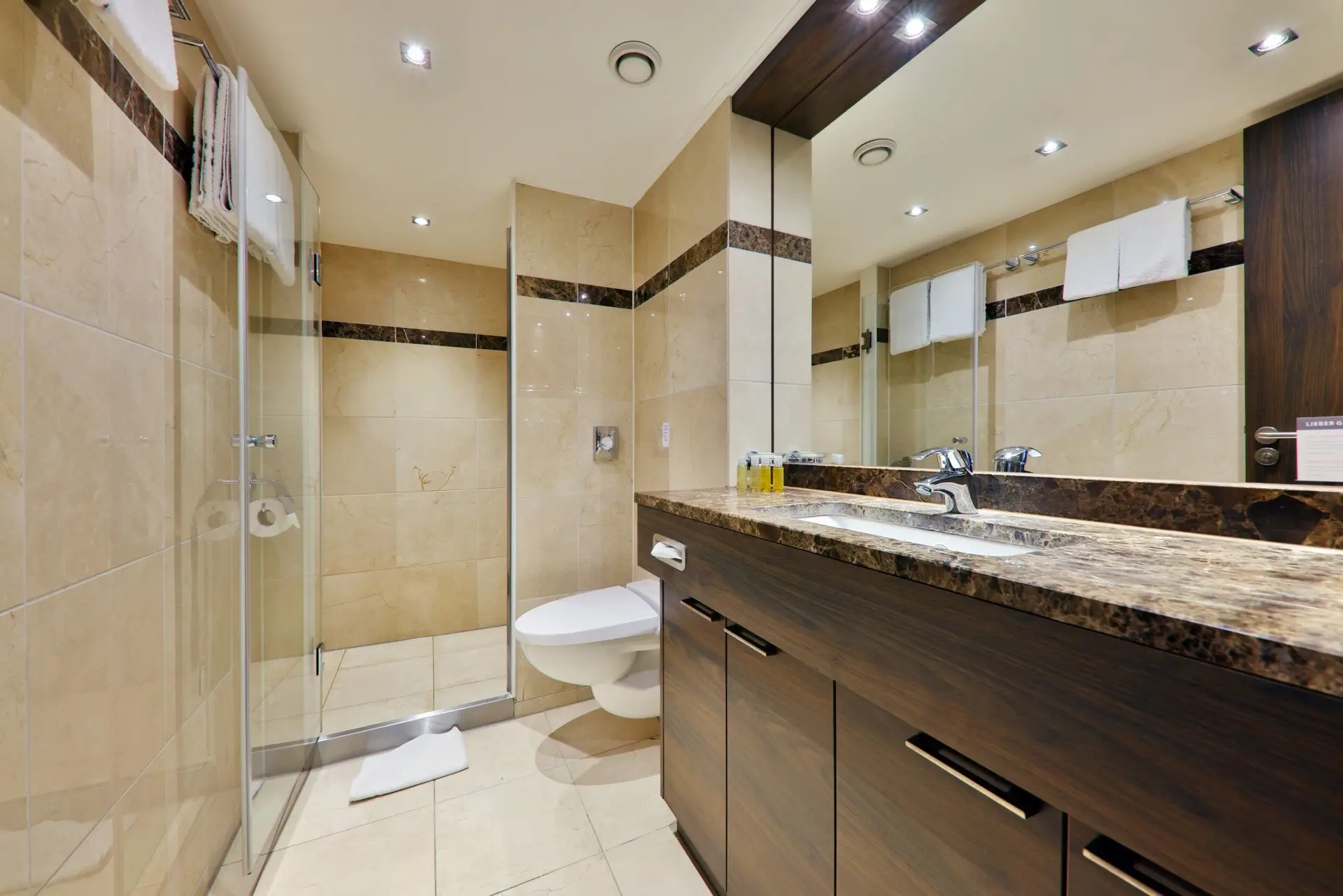 Badezimmer mit beiger Fliesenwand, Glasdusche, wandhängendem WC und dunklem Waschtisch mit großem Spiegel und zwei Wasserhähnen