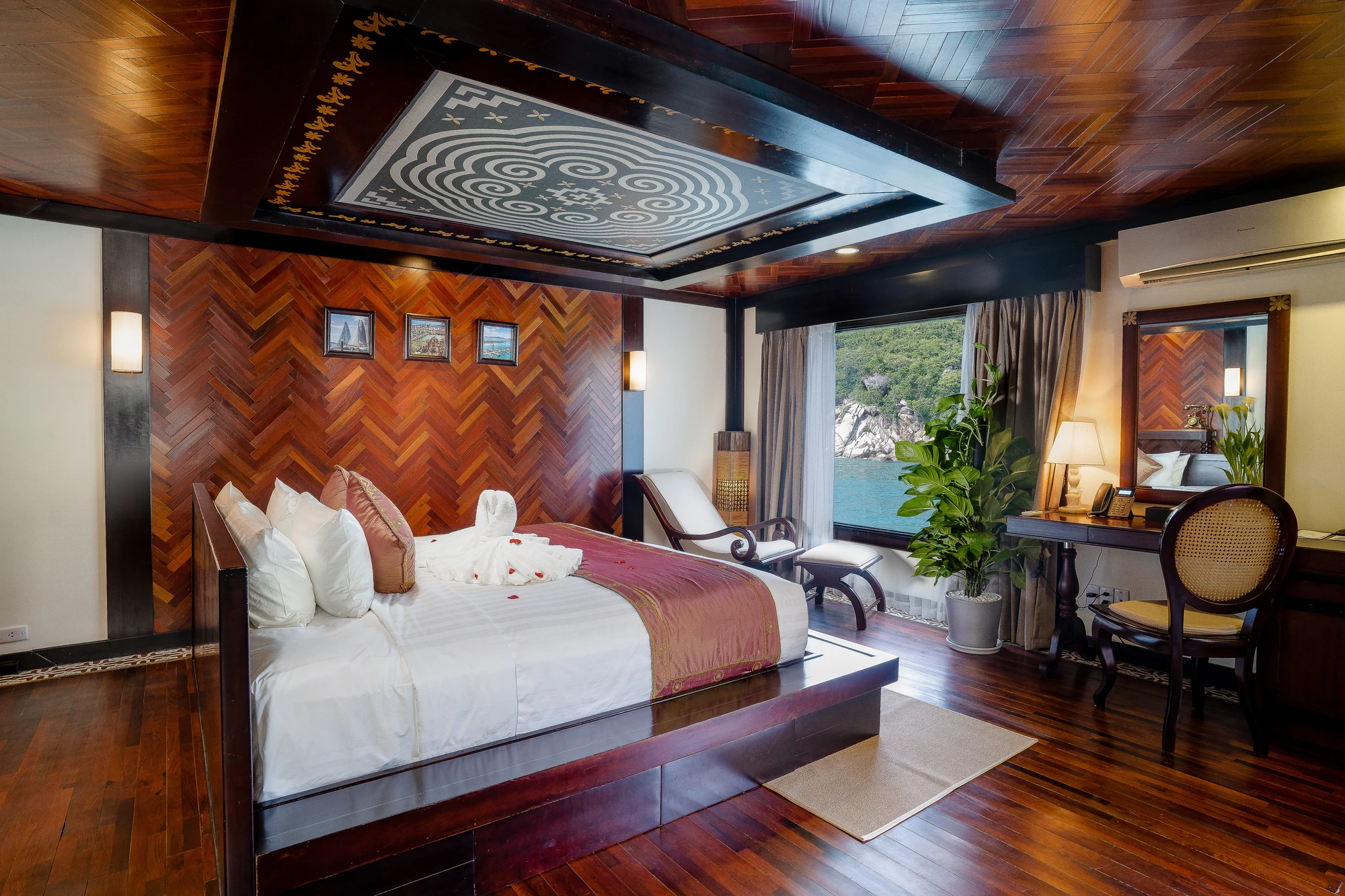 Luxuriöses Schlafzimmer mit einem großen Bett, Holzfußboden und hölzernem Wanddesign. Ein Schreibtisch mit Stuhl und Pflanzen sind sichtbar, sowie ein Fenster mit Blick auf das Wasser.