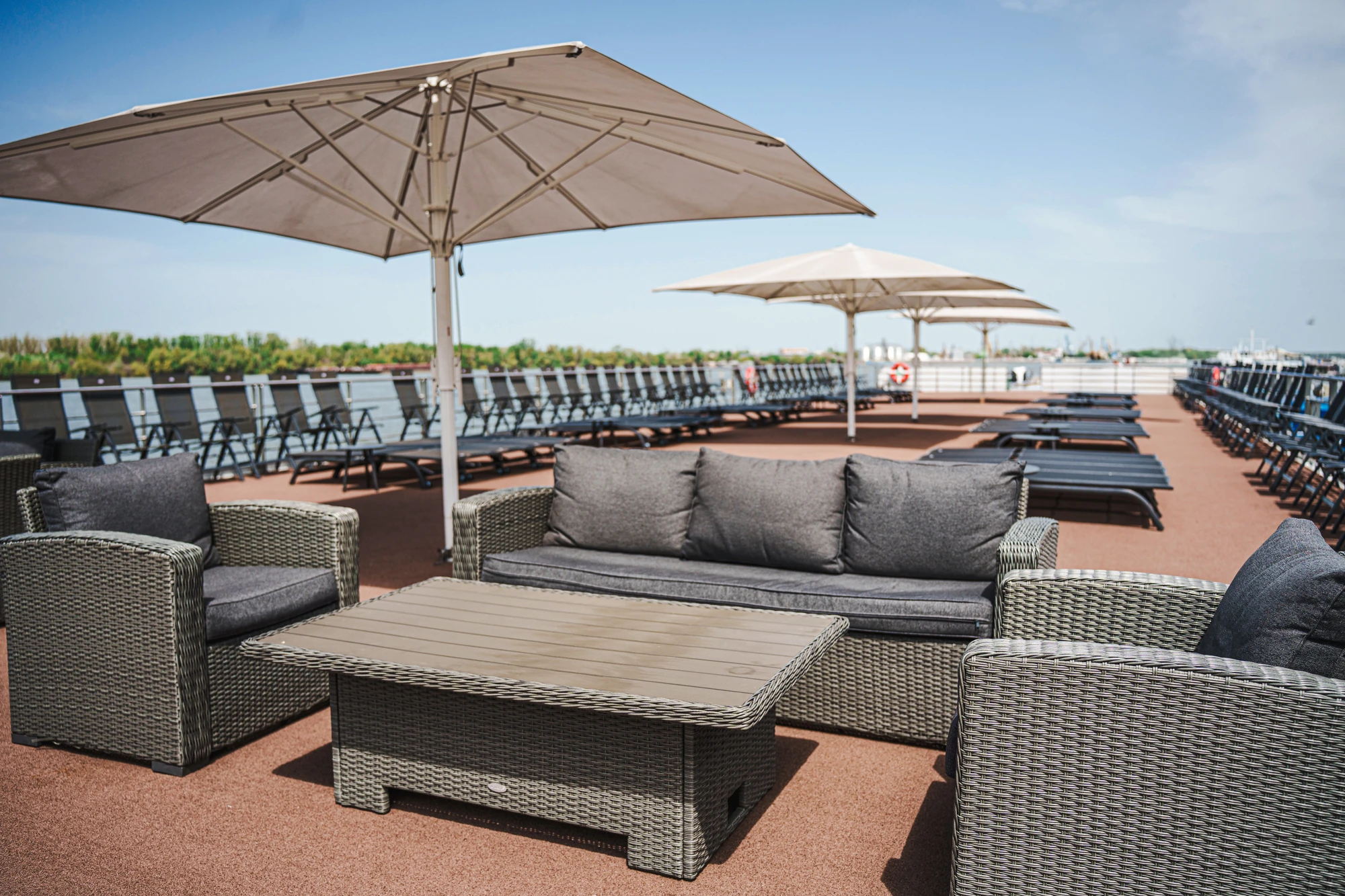 Sitzgruppe mit grauen Polstermöbeln und großen Sonnenschirmen auf einem Sonnendeck mit Reihen von Liegestühlen im Hintergrund