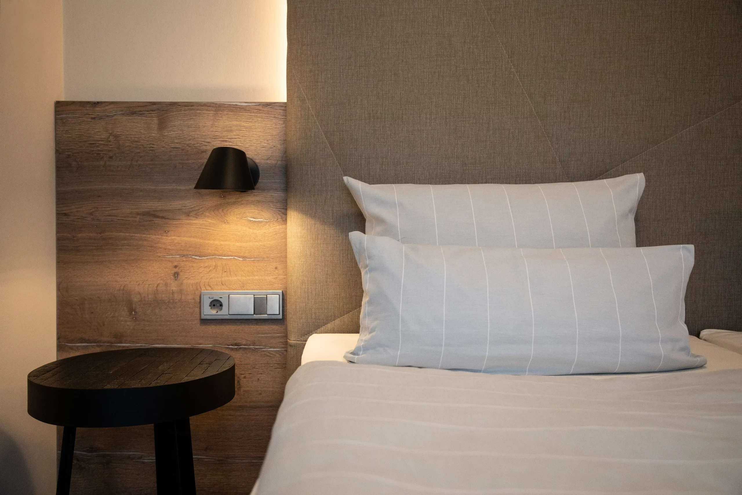 Schlafzimmer mit weiß gestreiften Kissen auf weißem Bett, Holzwand mit schwarzer Wandlampe und rundem schwarzen Nachttisch