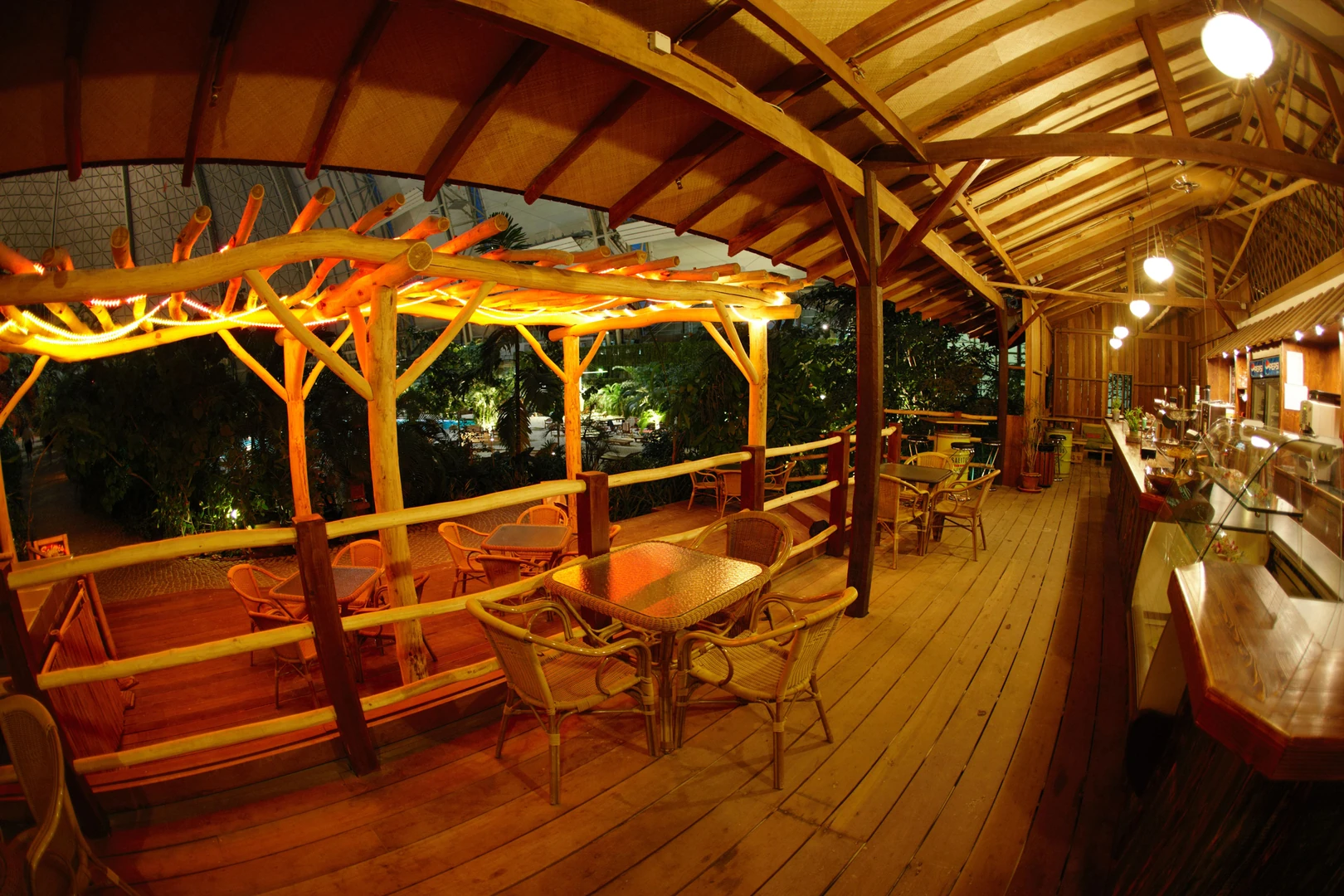 Das Bild zeigt eine tropische Bar und Restaurantterrasse im Tropical Islands, gestaltet im rustikalen Stil mit Holzboden, Bambusmöbeln und einer offenen Bauweise. Die stimmungsvolle Beleuchtung mit warmen Lichtern unterstreicht das entspannte Urlaubsambiente. Die Sitzbereiche bieten einen erhöhten Blick auf die üppige tropische Vegetation und das weitläufige Badeparadies. Die Kombination aus natürlichem Holz, gemütlichen Rattanmöbeln und exotischer Atmosphäre lädt zum Verweilen ein.