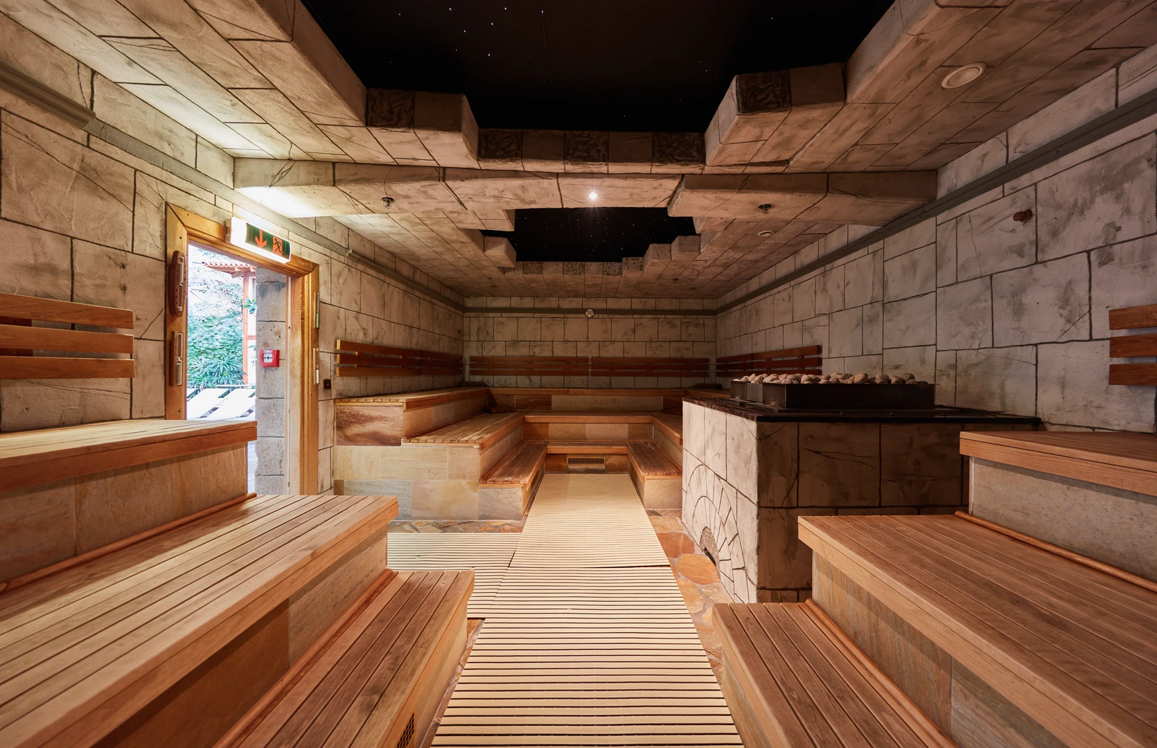 Das Bild zeigt die Steinsauna im Tropical Islands, inspiriert von antiker Architektur. Die Wände und Decke sind mit Steinoptik gestaltet, während die Sitzbänke aus warmem Holz bestehen. Im Zentrum befindet sich ein großer Saunaofen mit Natursteinen. Eine besondere Atmosphäre entsteht durch die dunkle Decke mit kleinen Lichtpunkten, die an einen Sternenhimmel erinnern. Die großzügige Bauweise und die dezente Beleuchtung schaffen eine entspannende und luxuriöse Umgebung.