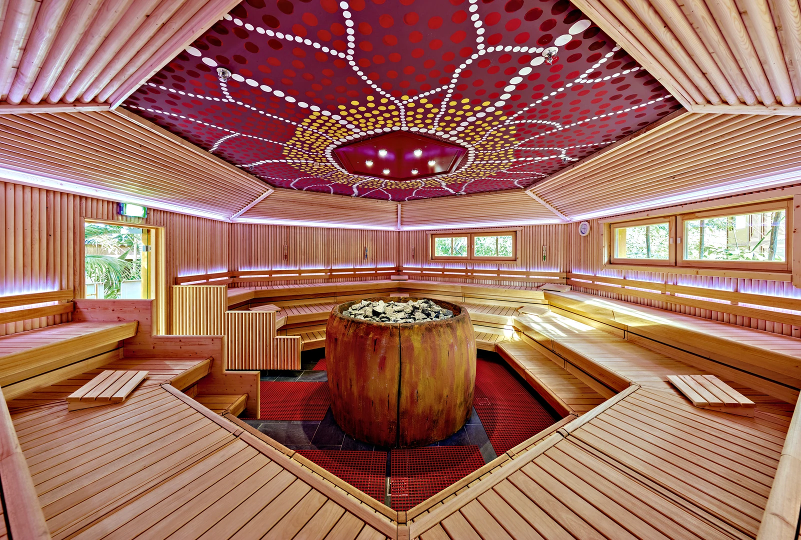 Das Bild zeigt eine großzügige Sauna im Tropical Islands, die mit hellem Holz und modernen Lichtelementen gestaltet ist. Im Zentrum befindet sich ein großer Saunaofen in einem rustikalen Steingefäß. Die Sitzbänke sind stufenförmig angeordnet und bieten viel Platz für Gäste. Die Decke ist kunstvoll mit einem bunten Muster im Stil australischer Aborigine-Kunst verziert. Große Fenster lassen Tageslicht herein und ermöglichen einen Blick auf die tropische Umgebung, wodurch eine entspannte und exotische Atmosphäre entsteht.