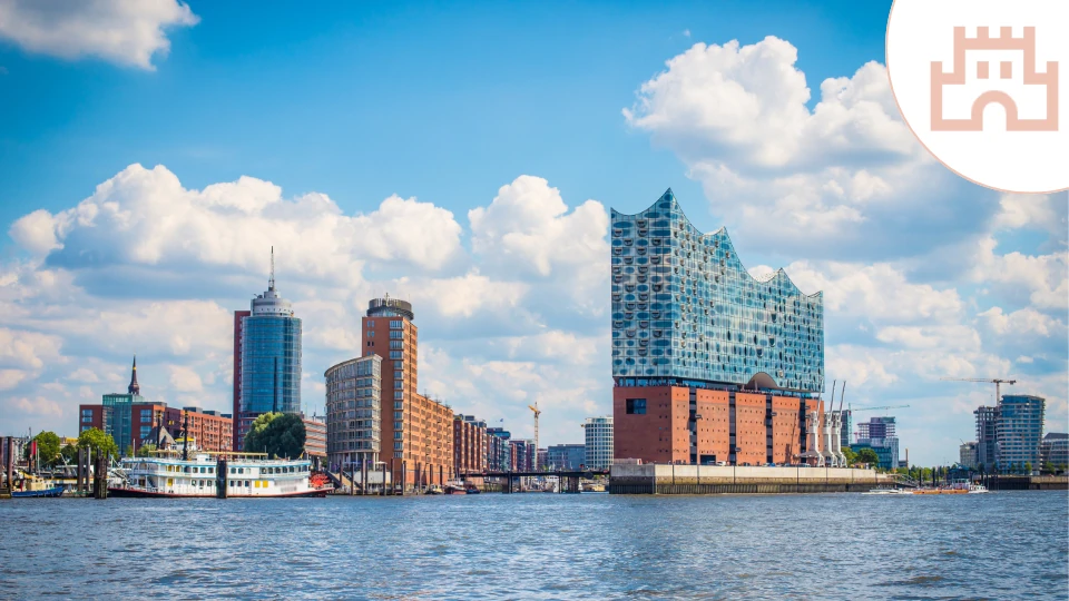 Das Bild zeigt die Hamburger Elbphilharmonie, eines der markantesten Wahrzeichen der Stadt, mit ihrer wellenförmigen Glasfassade über dem historischen Kaispeicher. Im Vordergrund fließt die Elbe, während ein klassisches Schiff am Ufer liegt. Die Skyline kombiniert moderne Hochhäuser mit traditioneller Hafenarchitektur unter einem Himmel mit weißen Wolken.