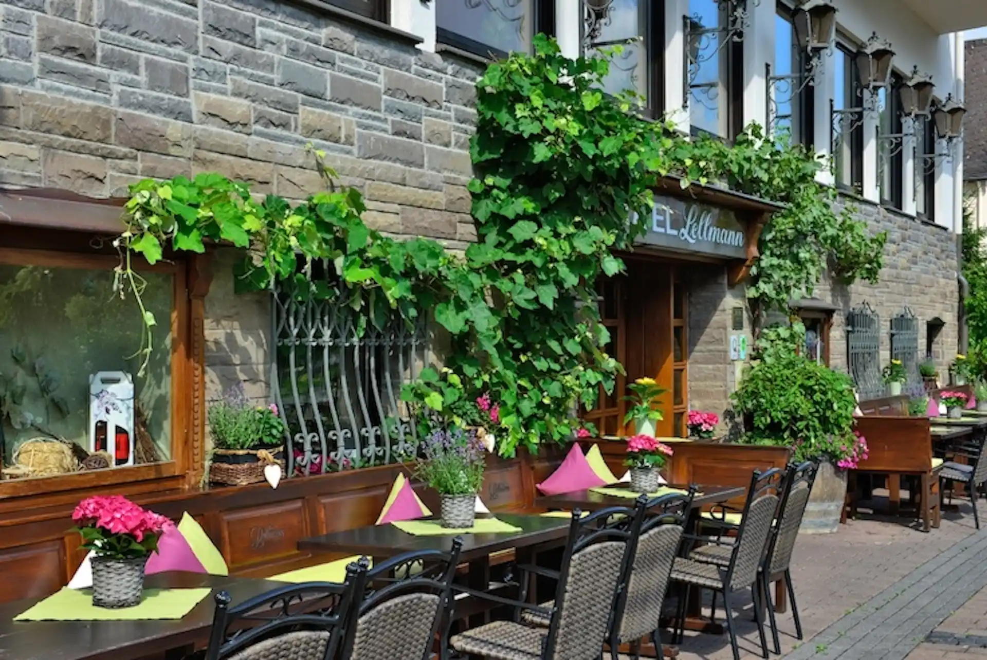 Terrassenbereich mit Holztischen, Stühlen, grünen Pflanzen und bunten Kissen vor einer Steinmauer mit Fenstern
