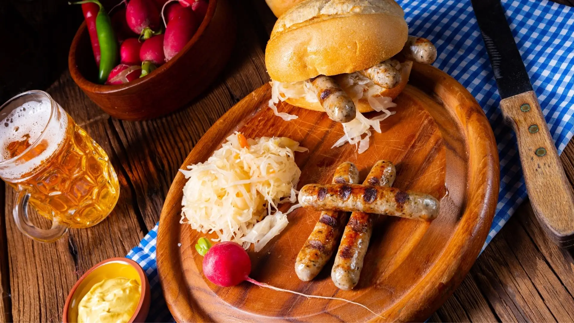 Drei gegrillte Thüringer Rostbratwürste auf Holzbrett mit Sauerkraut, Radieschen, Brötchen, Senf, Bier und Schale mit roten und grünen Chilischoten.