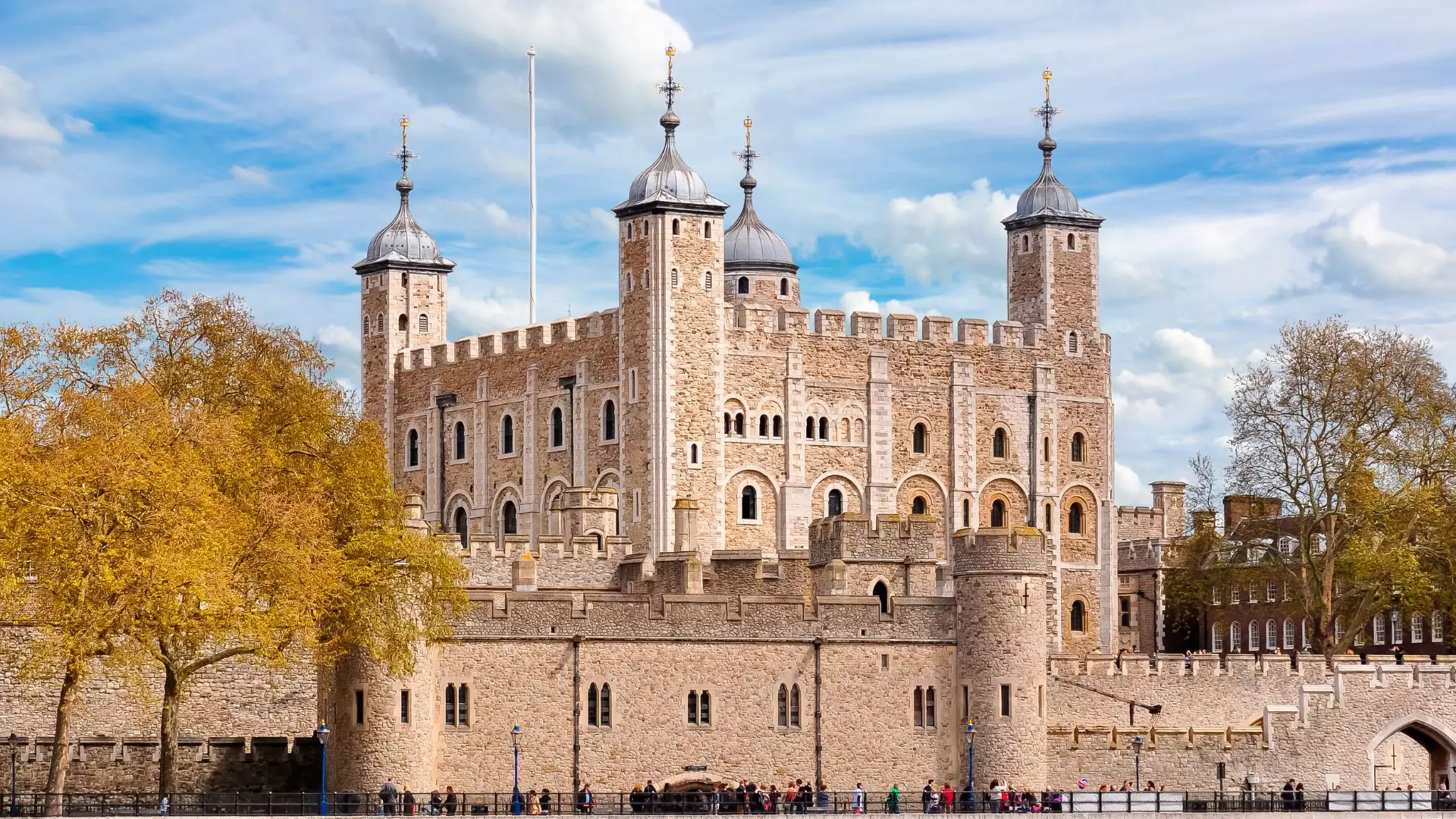 Blick auf den Tower of London mit seinen vier runden Ecktürmen und Zinnen, umgeben von einer Steinmauer und Bäumen unter blauem Himmel.