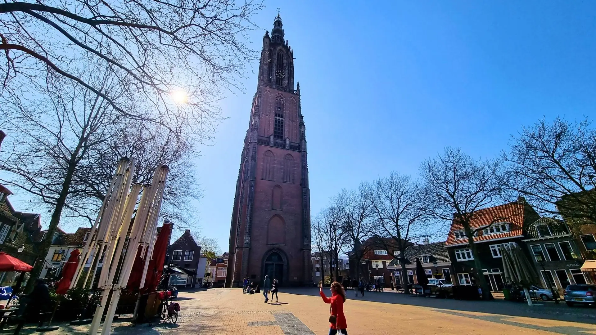 Hoher, schlanker Backsteinturm der Onze-Lieve-Vrouwetoren in Amersfoort bei klarem Himmel, umgeben von Bäumen und historischen Gebäuden.