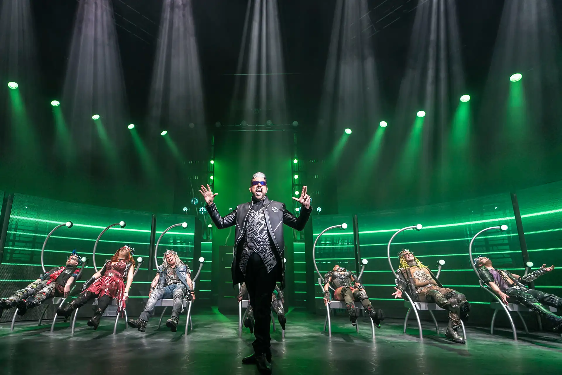 We will Rock You – Das Musical. Szenenbild: Darsteller mit Sonnenbrille im Vordergrund, weitere Darsteller in Stühlen im Hintergrund. (UK Cast)