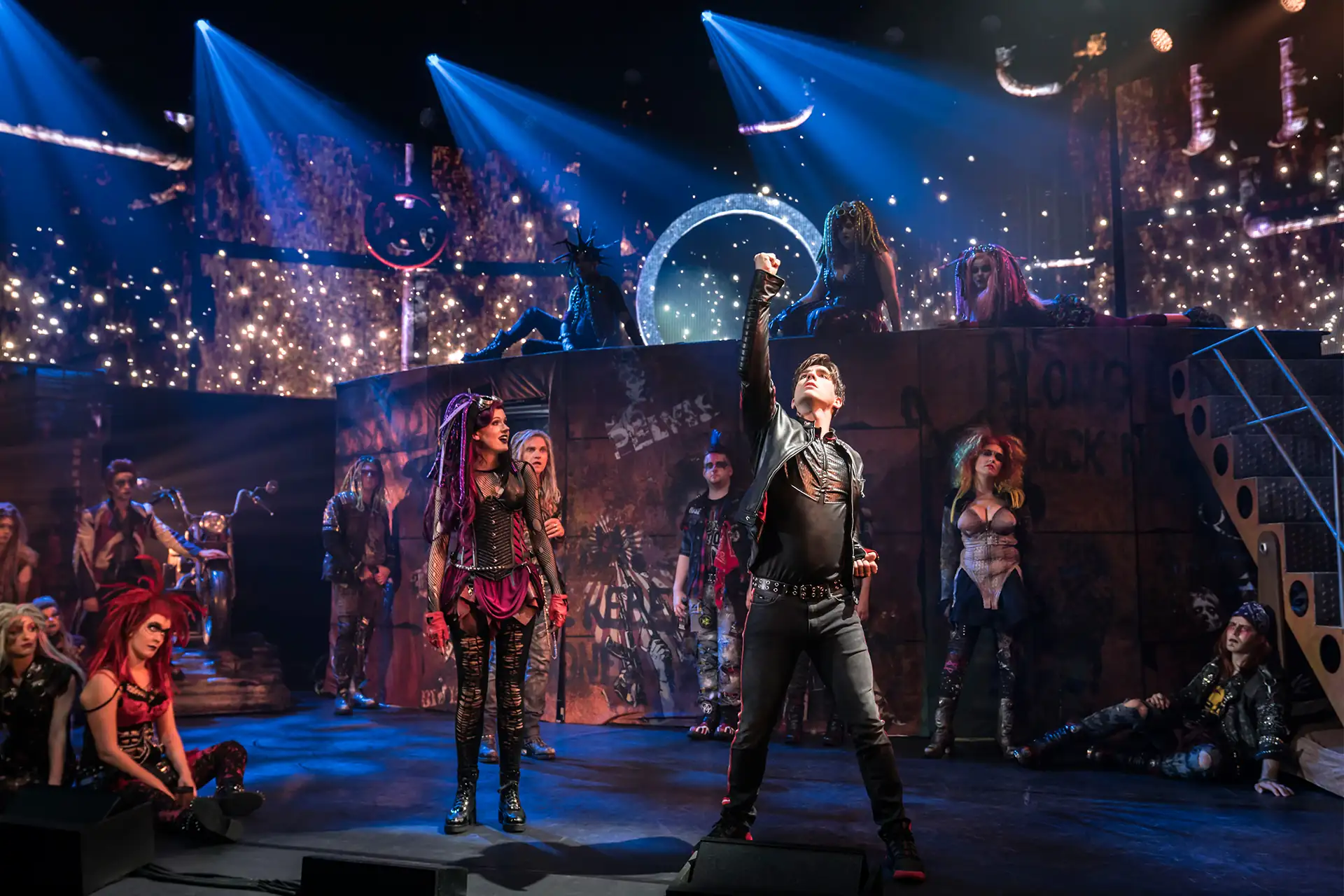 We will Rock You – Das Musical. Szenenbild: Darsteller in schwarzer Kleidung reckt die Faust in die Höhe im dystopischen Setting. (UK Cast)