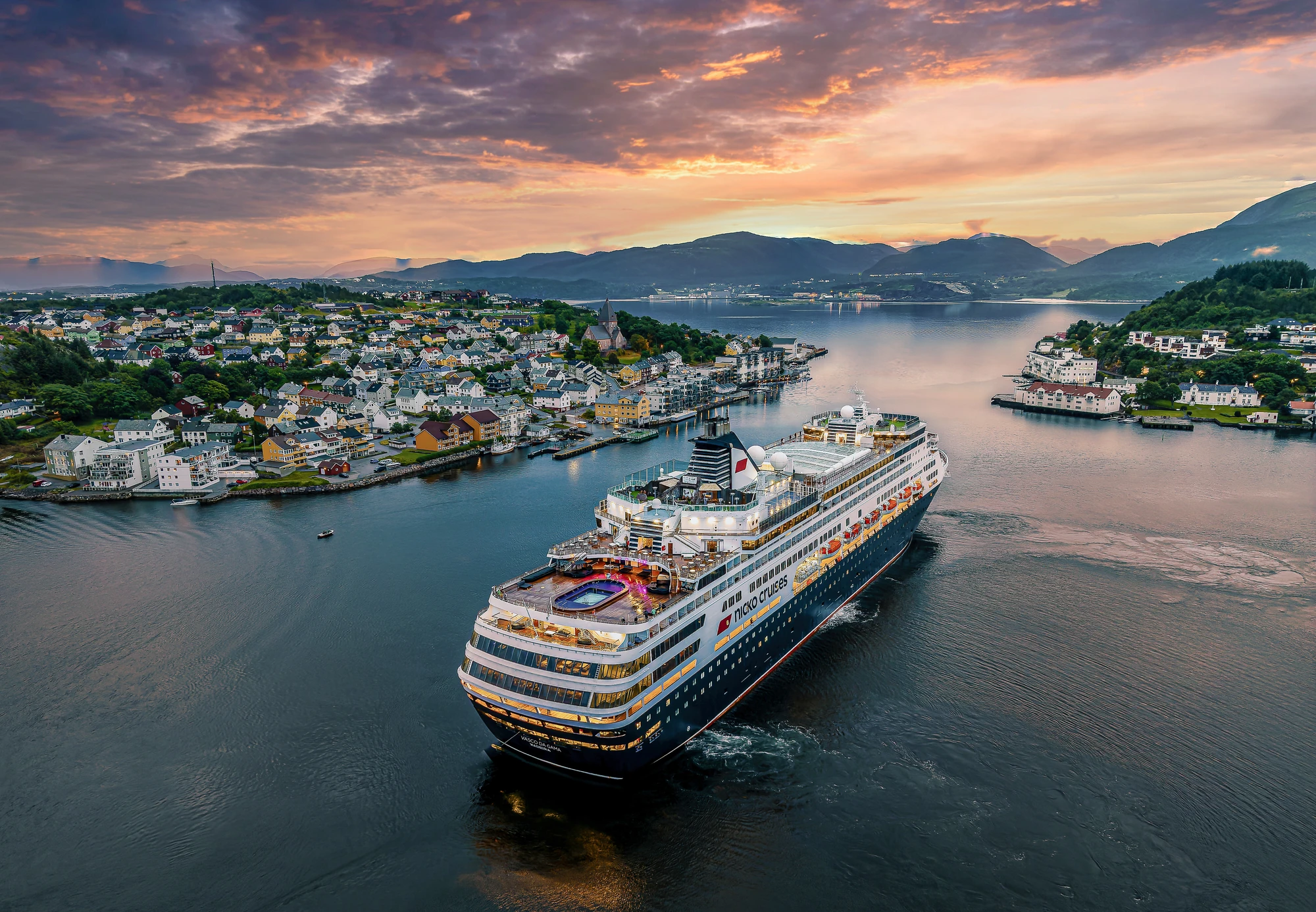 Ein großes Kreuzfahrtschiff fährt durch eine ruhige Fjordlandschaft mit Häusern an der Küste und Bergen im Hintergrund. Der Himmel ist bei Sonnenuntergang in warmen Farben gefärbt.