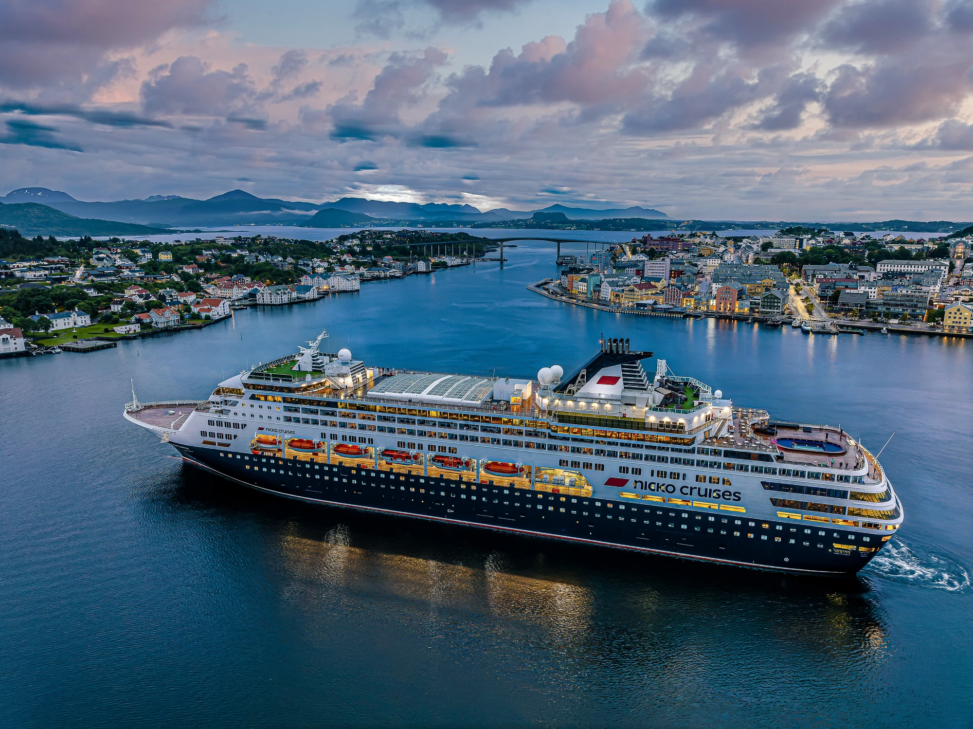 Ein Kreuzfahrtschiff der Marke Nicko Cruises mit orangefarbenen Lifeboats fährt durch eine ruhige Wasserstraße, umgeben von einer Stadt und Bergen im Hintergrund.