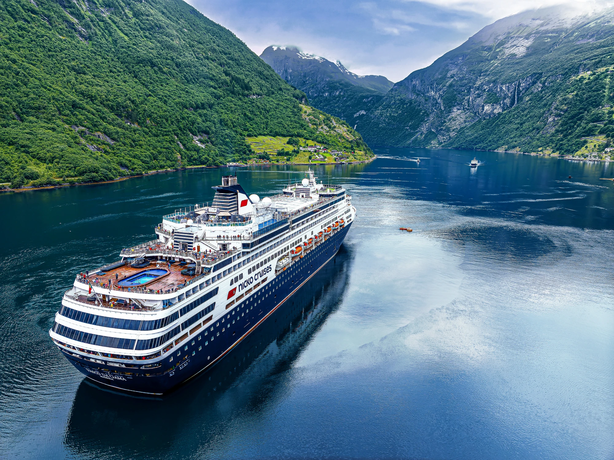 Ein großes Kreuzfahrtschiff mit blauer und weißer Farbe liegt im Geirangerfjord, umgeben von grünen Bergen und ruhigem Wasser.
