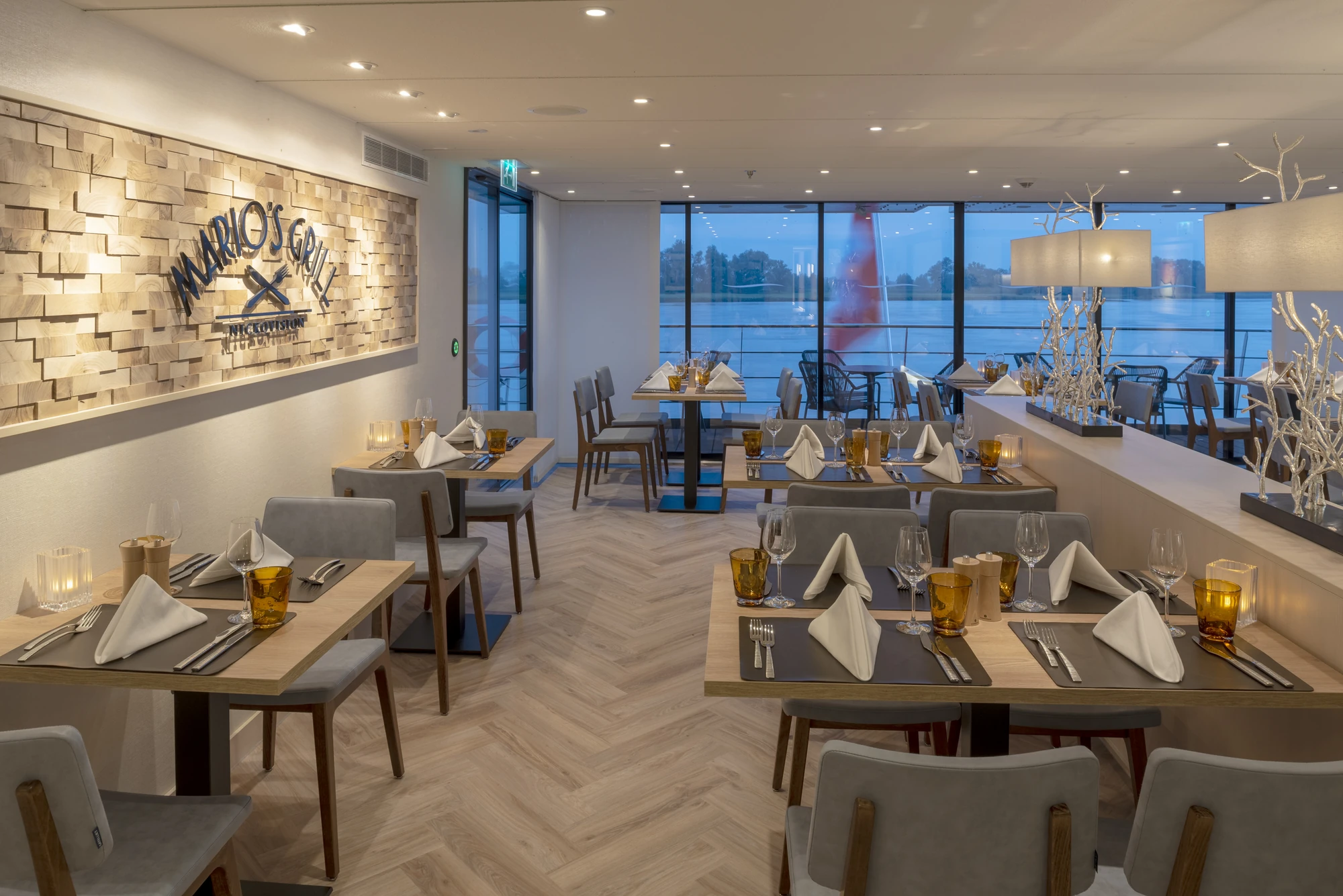 Innenansicht des Restaurants 'Marios Grill' auf der nickoVISION, mit Holzwand, Tischen mit gedeckten Tellern und Stühlen. Große Fenster mit Blick auf Wasser.