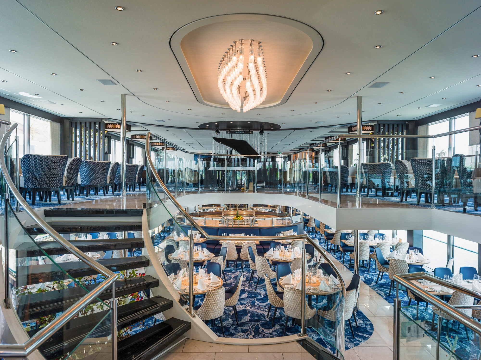 Innenansicht des Panorama Salons auf der nickoVISION mit zwei Etagen, geschwungener Treppe und blauen Stühlen. Ein Kronleuchter hängt von der Decke.