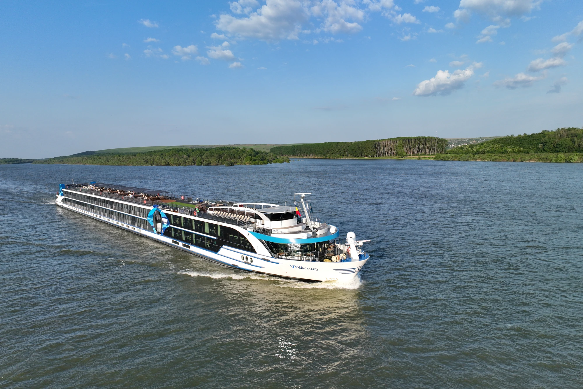 Flusskreuzfahrtschiff auf dem Donaufluss, umgeben von bewaldeten Uferbereichen unter blauem Himmel mit vereinzelten Wolken.