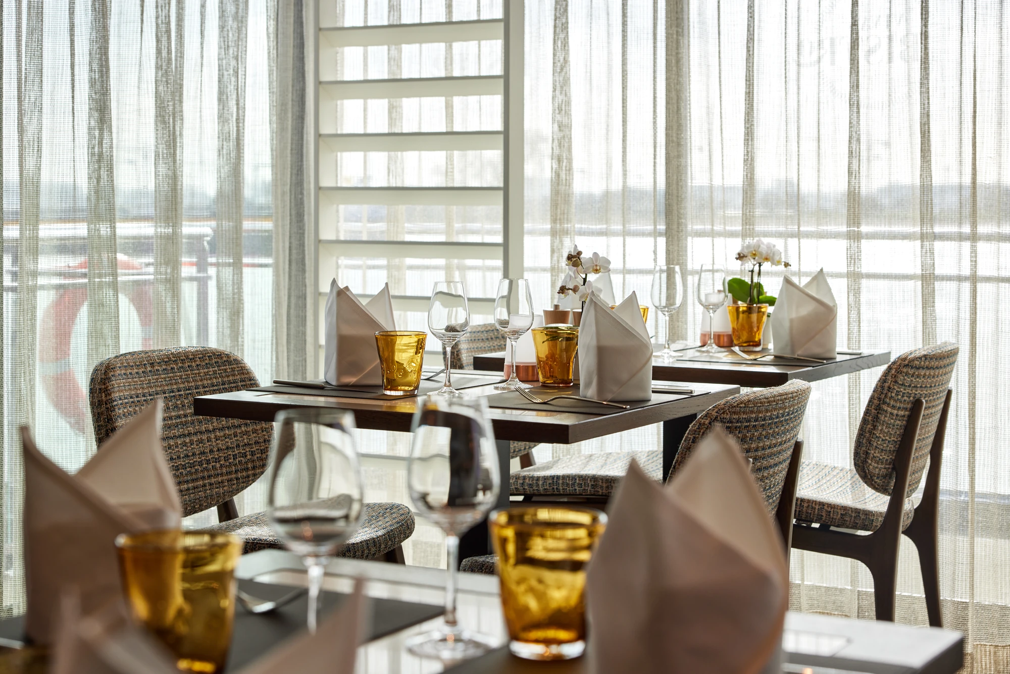 Bistro-Tisch mit gefalteten weißen Servietten, gelben Gläsern, Weingläsern und gepolsterten Stühlen vor großen Fenstern mit halbtransparenten Vorhängen.