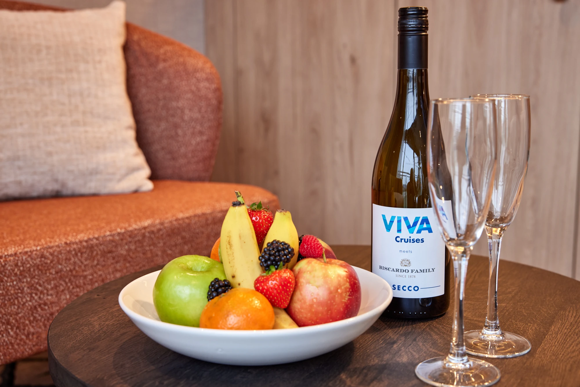 Weißer Teller mit grünem Apfel, rotem Apfel, Banane, Mandarine, Erdbeeren und Brombeeren auf braunem Tisch neben Flasche VIVA Cruises Secco und zwei leeren Sektgläsern, im Hintergrund braunes Sofa mit Kissen.