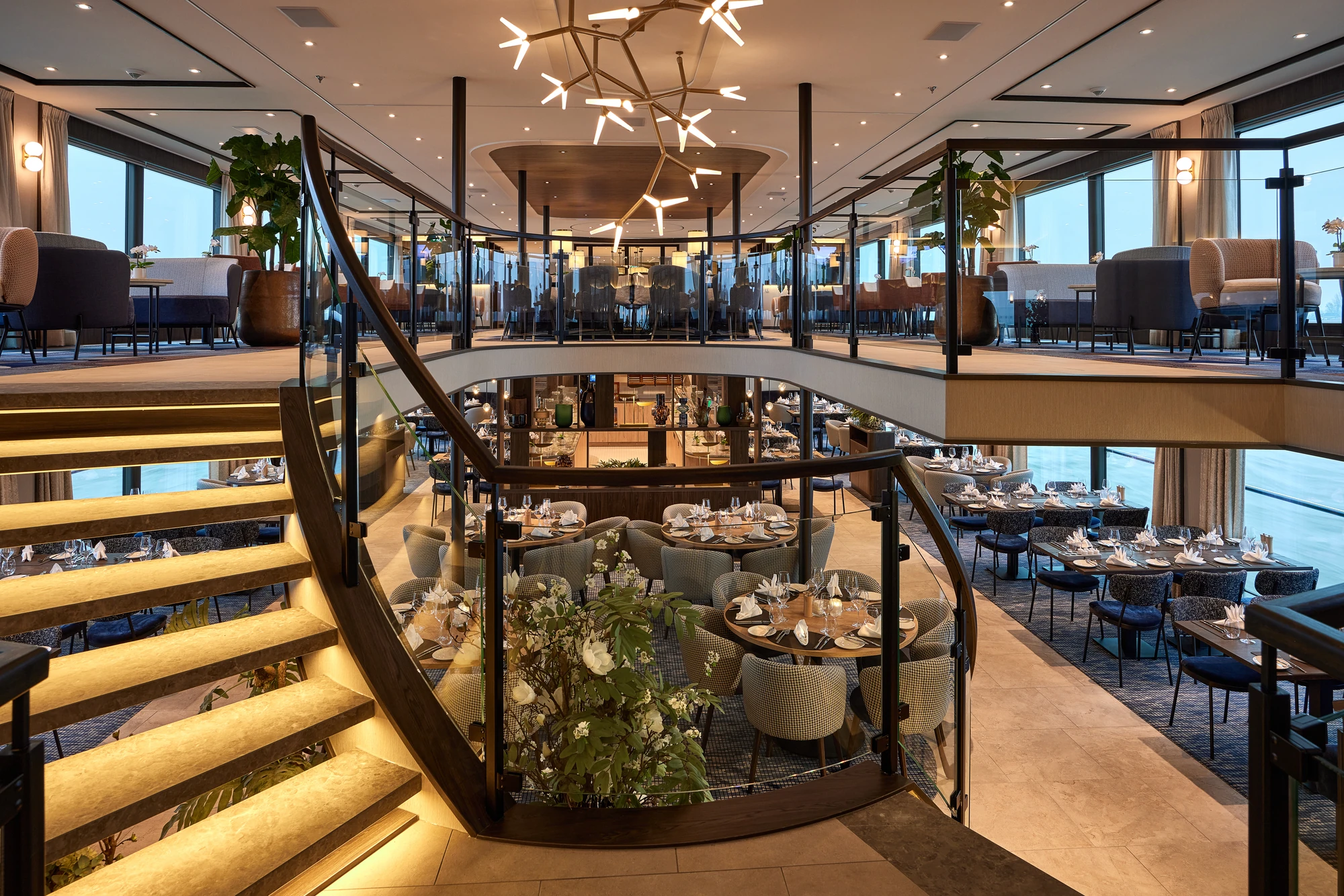 Zweistöckige Lounge mit modernen Sitzgruppen, runden Tischen, Glasgeländer und beleuchteter Treppe, große Fenster mit Meerblick.