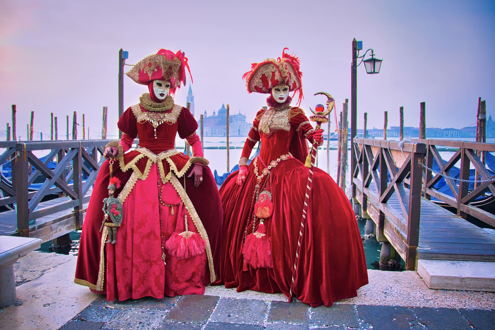 Zwei Gäste im roten Kostüm und mit kunstvollen Masken stehen beim Karneval in Venedig vor der malerischen Kulisse der Stadt. Die sanfte Abenddämmerung taucht die Umgebung in warmes Licht und verstärkt die festliche Atmosphäre, während die prachtvollen Kostüme und Masken im Mittelpunkt stehen.