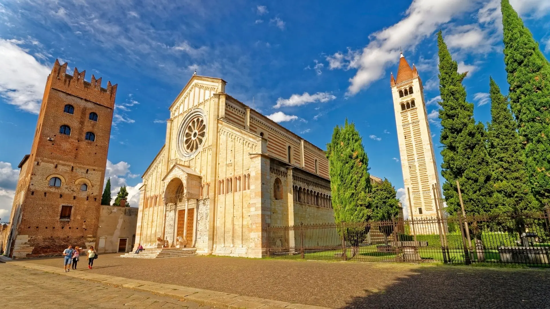 Basilika San Zeno Maggiore in Verona mit romanischer Fassade, Rosettenfenster, zwei Türmen und Zypressen unter blauem Himmel.